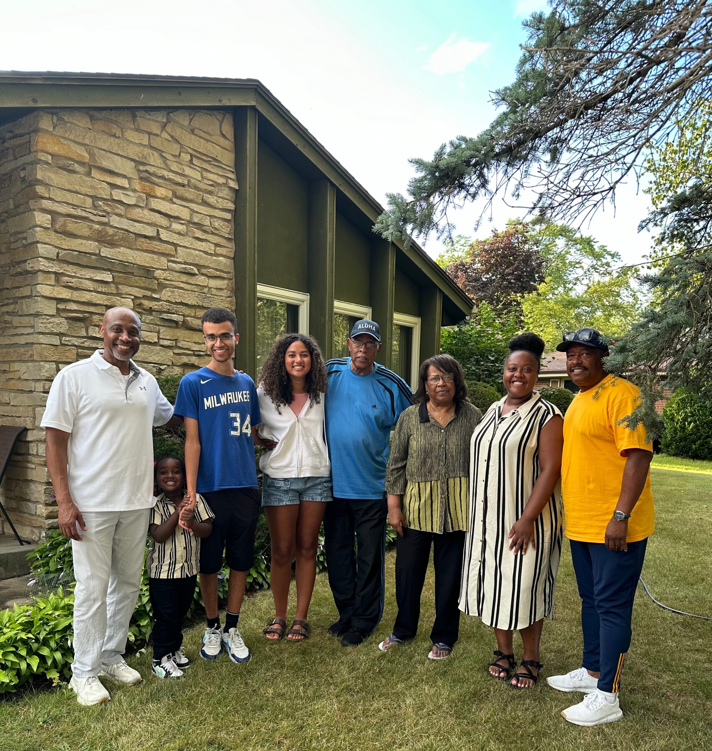 Group of people posing together in a front yard beside a stone house, smiling outdoors.
