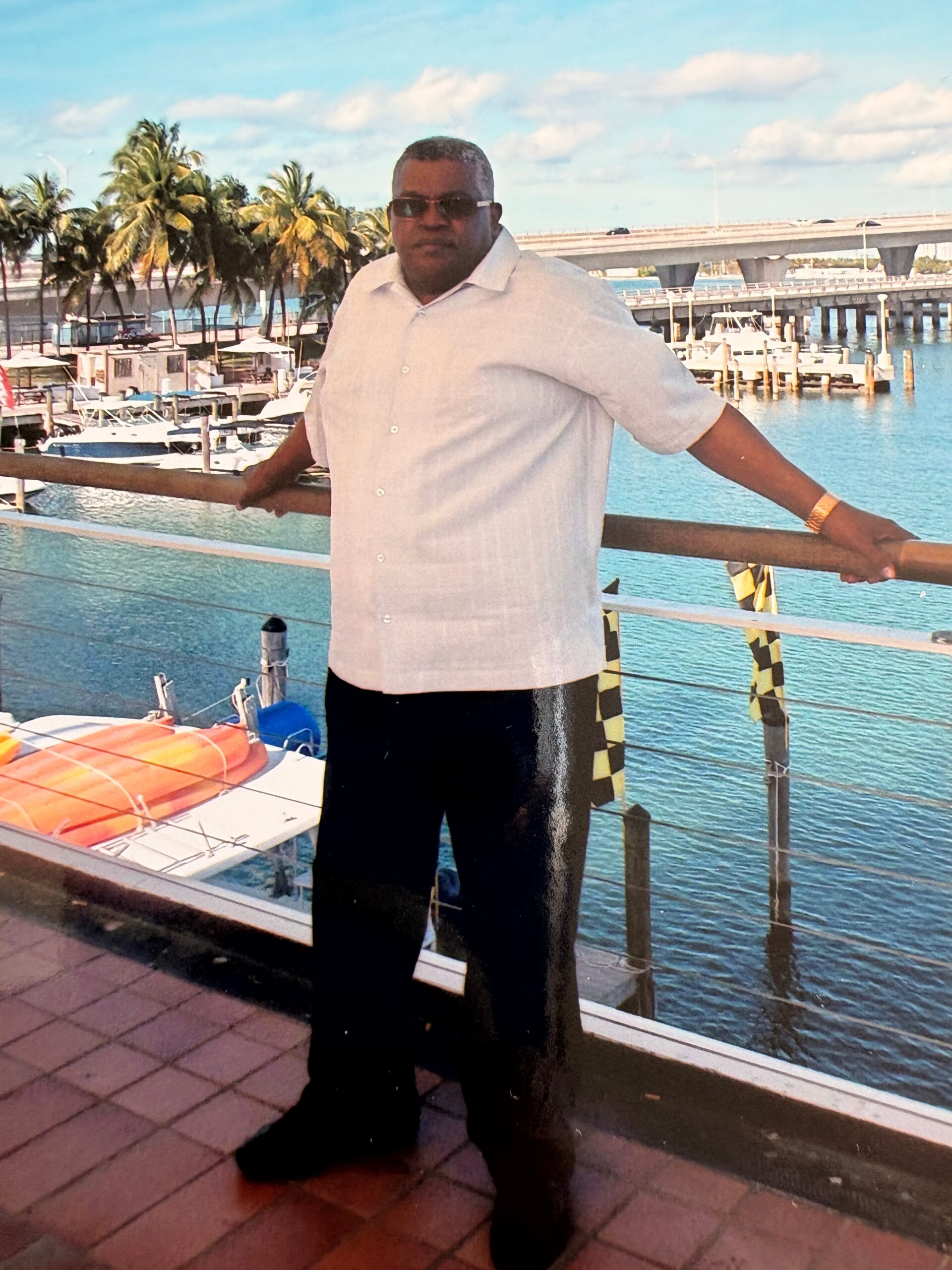 Man in a light shirt and sunglasses leaning on a railing at a marina with boats and palm trees in the background.