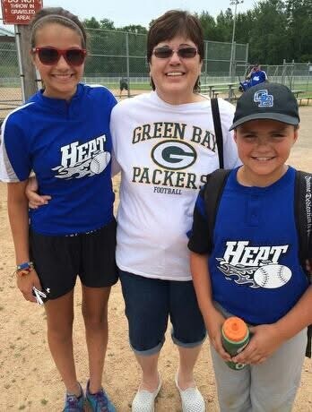 Three people pose on a baseball/softball field: two girls in blue HEAT shirts flank a woman in a Green Bay Packers shirt, all smiling at the camera.