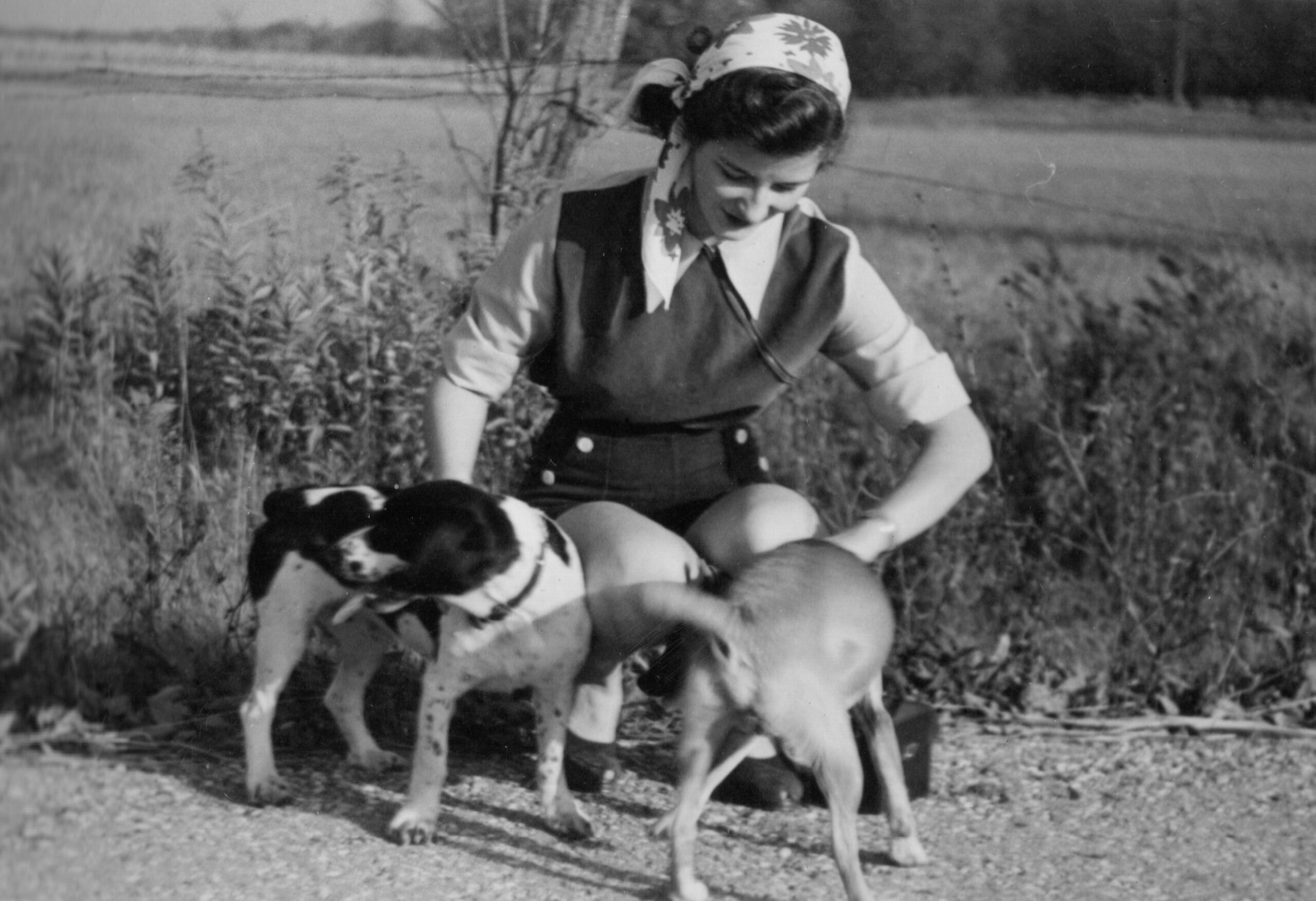 Young woman wearing a headscarf, vest and shorts kneels in a field, leaning toward a dog beside her.