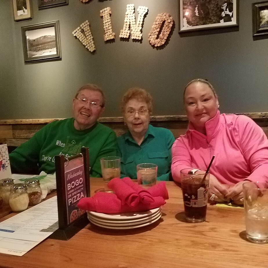 Three people sit at a wooden restaurant table, smiling at the camera with drinks and napkins in front; a cork-letter sign spelling VINO on the wall behind them.