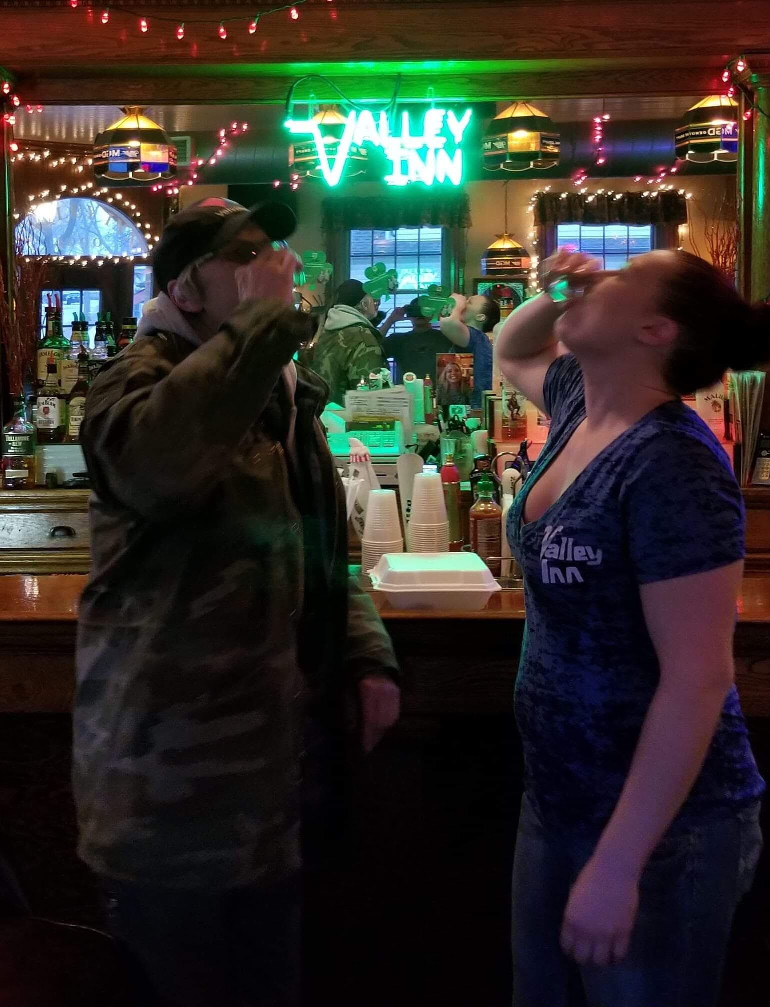 Two people at a bar toasting with raised glasses; a bright neon 'Valley Inn' sign and festive lights in the background show a lively bar scene.