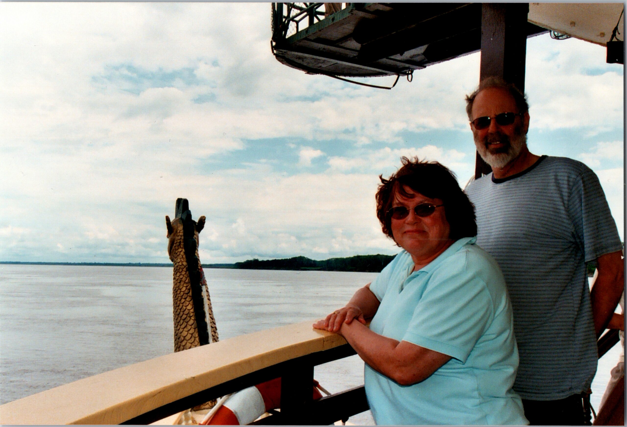 Two seniors with sunglasses leaning on a boat railing, posed by calm water and cloudy sky, with a carved figurehead nearby.
