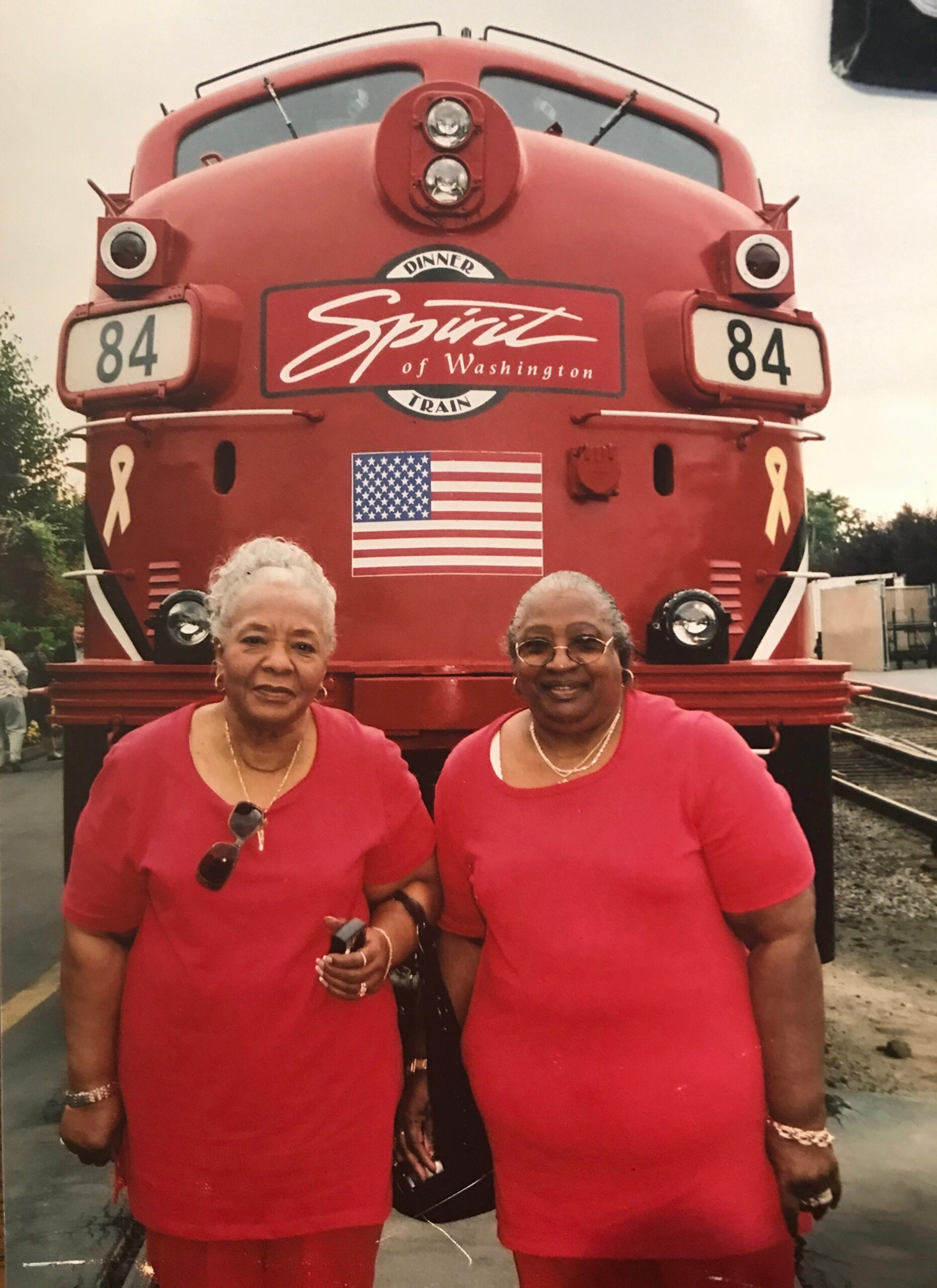 Two elderly women in red outfits pose in front of a red Spirit of Washington train with an American flag visible behind them.