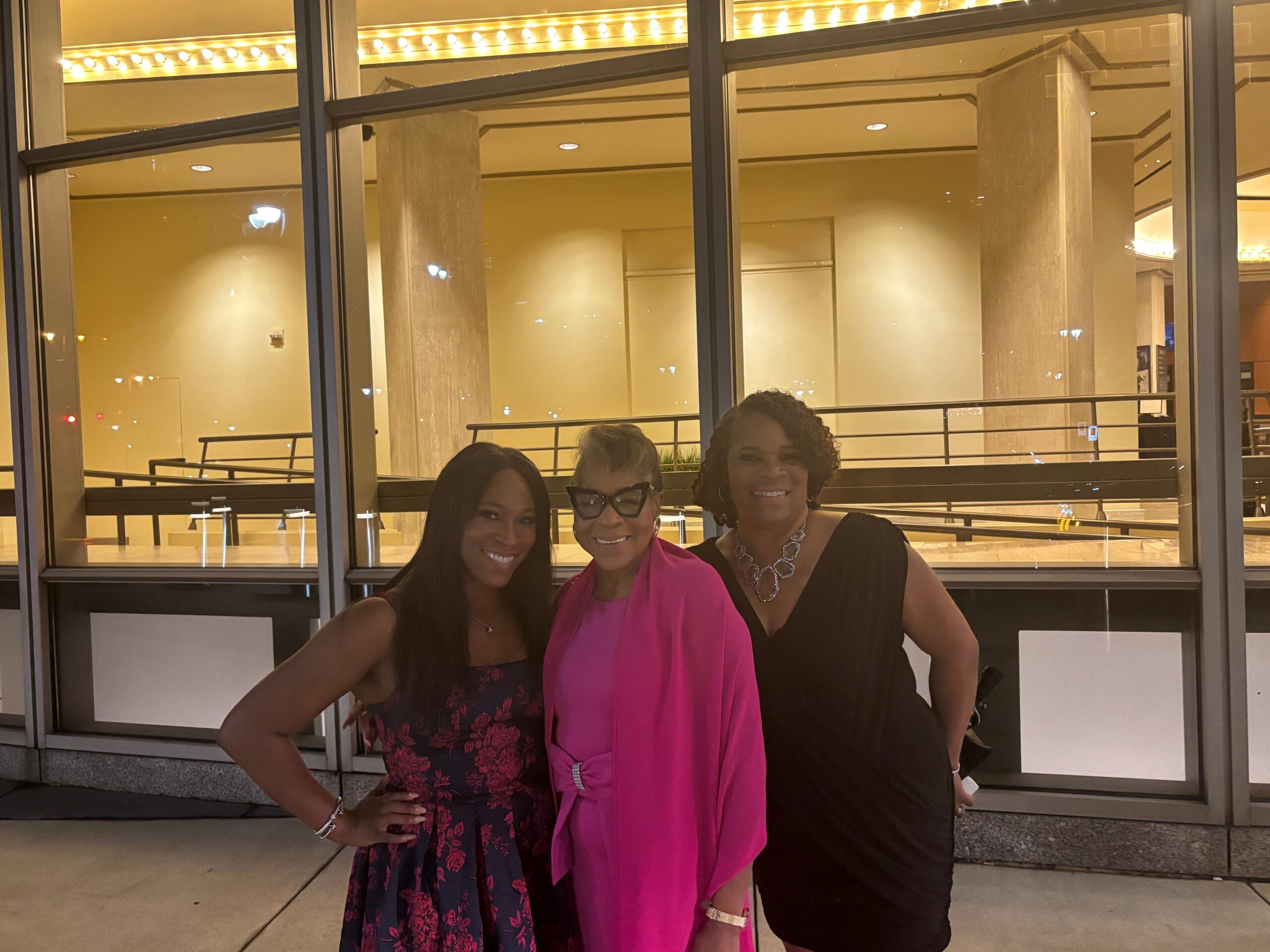 Three women in evening wear posing together outdoors in front of a glass storefront at night, smiling.