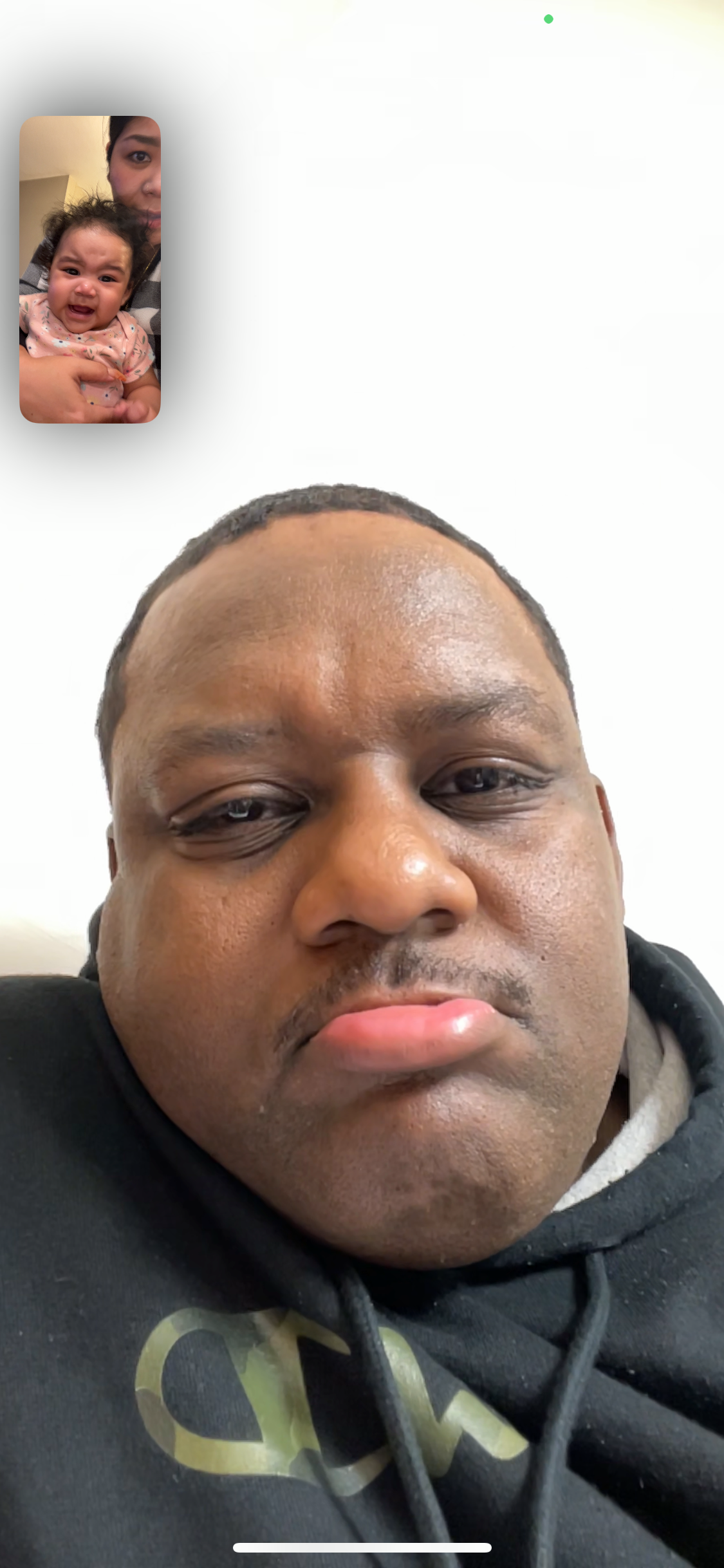 Man in a black hoodie taking a close-up selfie during a video call; small inset shows a crying baby being held by someone in the background