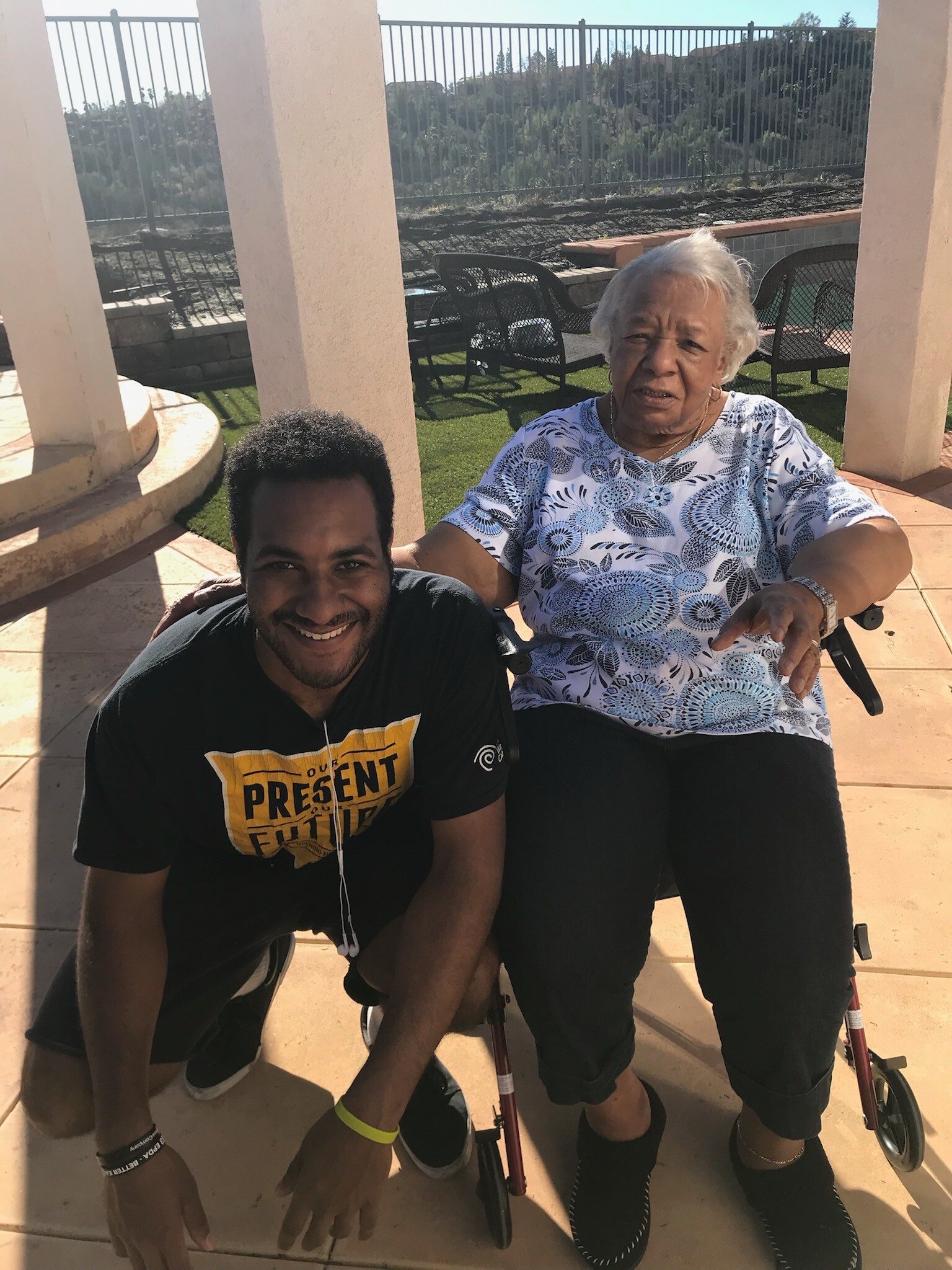 Smiling young man kneels beside an elderly woman in a wheelchair on a sunny patio, with a metal fence and pool area in the background.