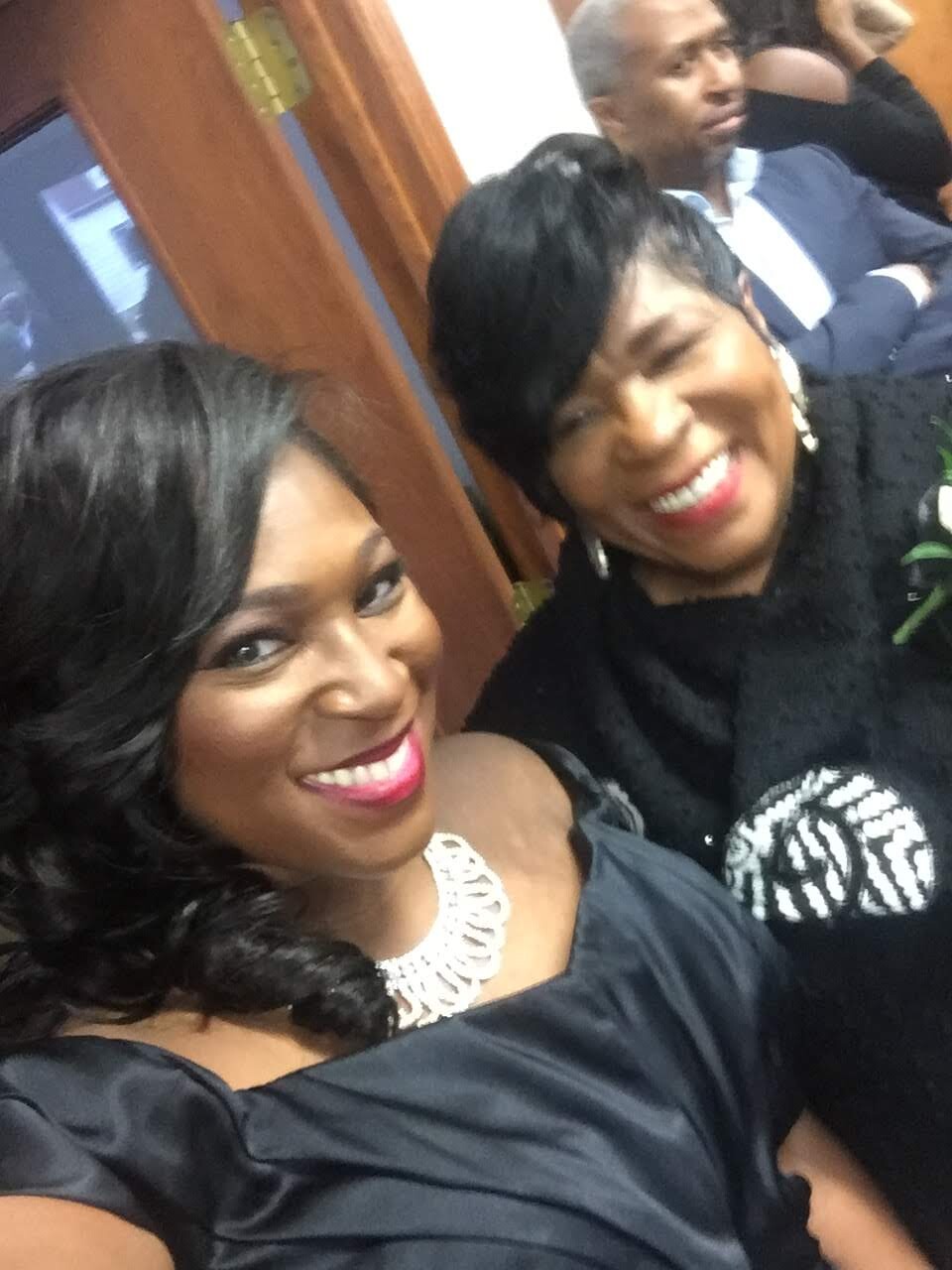 Two women in formal dresses smiling for a selfie at an event, with a man in a suit in the background.
