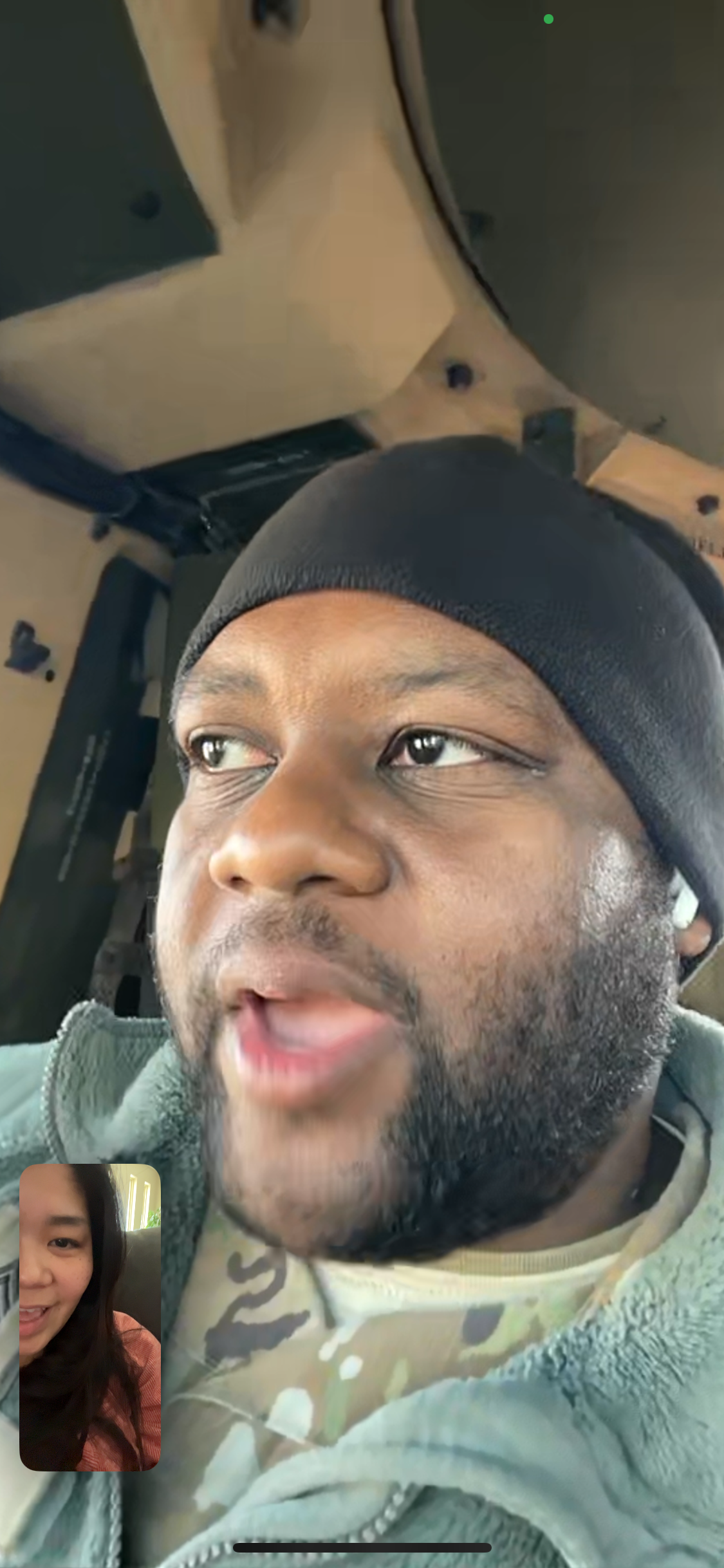 Man wearing a black beanie and camo jacket speaks during a video call, with a small inset video of a woman at the bottom left.
