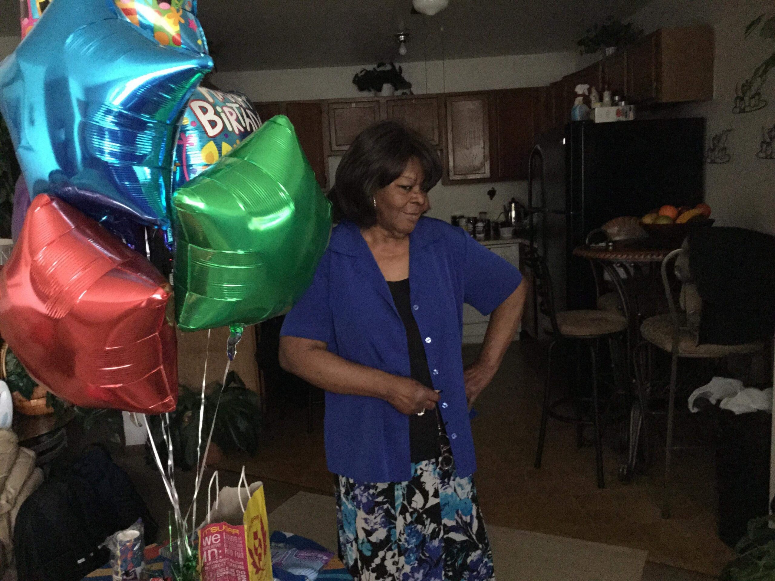 Woman in a blue shirt standing indoors with colorful birthday balloons and gifts nearby in a home setting.