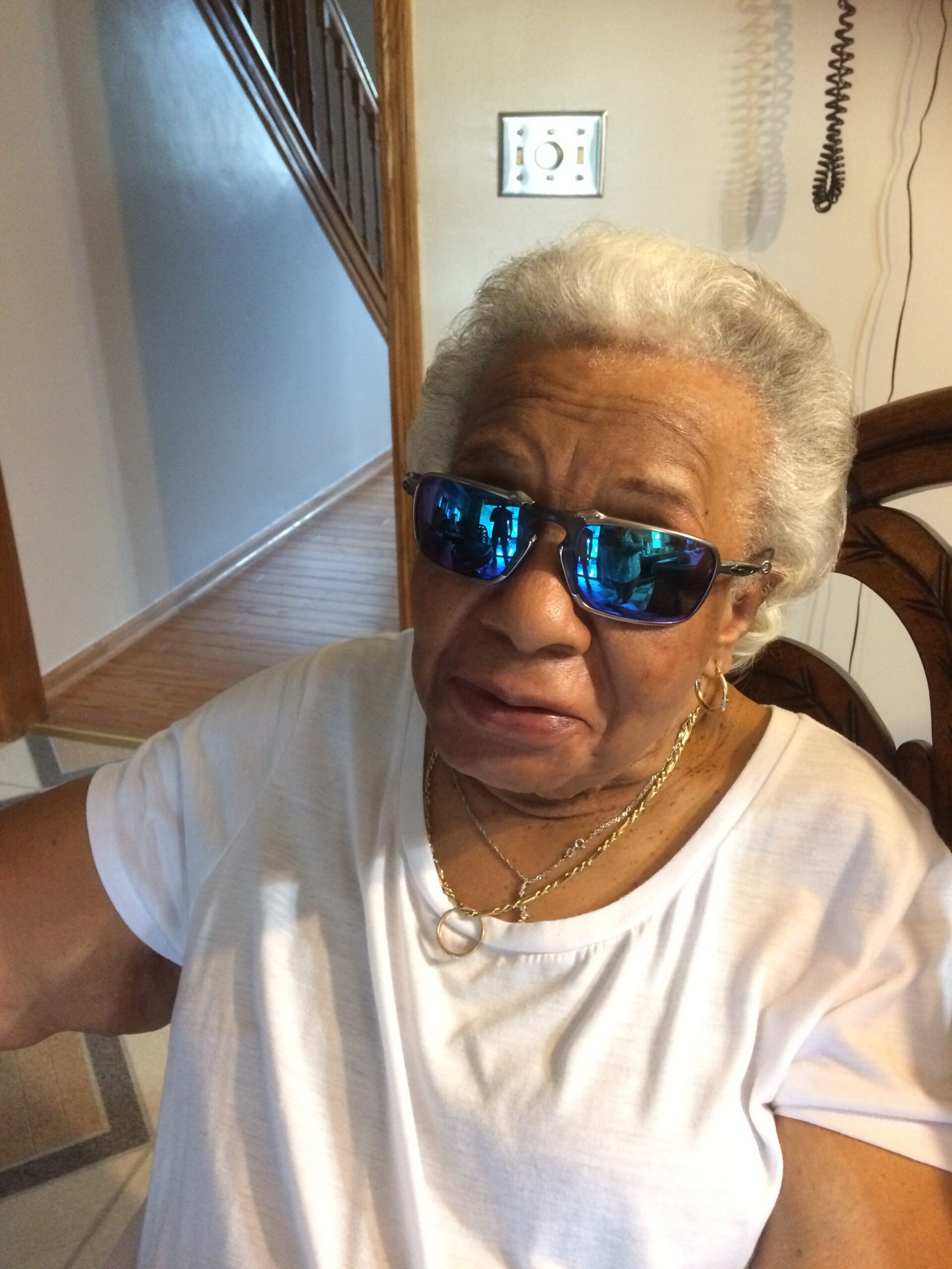 Close-up of an elderly woman wearing blue mirrored sunglasses and a white shirt, indoors, with gold jewelry