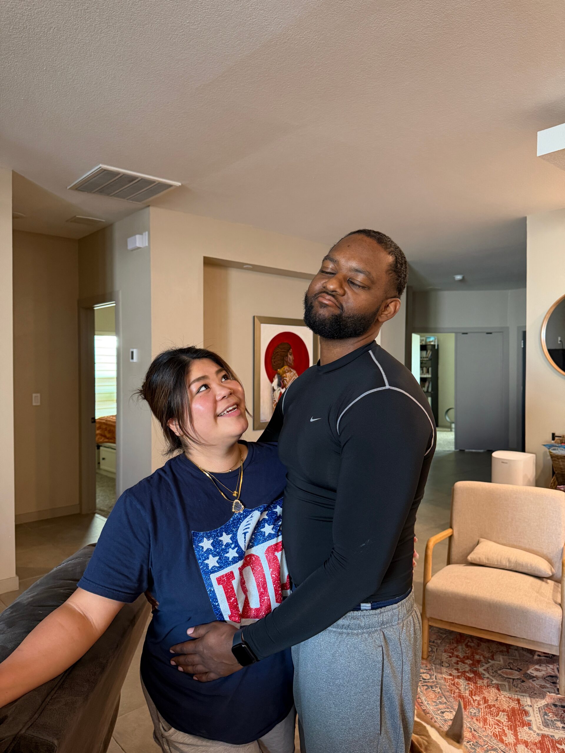 Woman in a blue graphic tee embraces a man in a black athletic shirt in a cozy living room, both smiling softly.