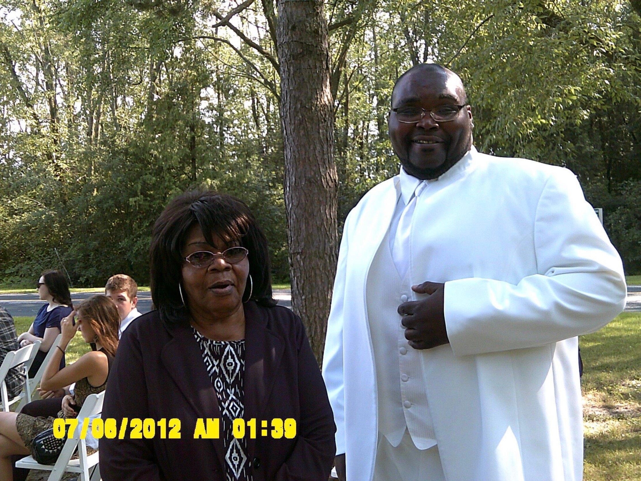 Smiling man in a white suit stands beside a seated woman with glasses at an outdoor event, with trees in the background and a yellow date/time stamp at the bottom of the image.