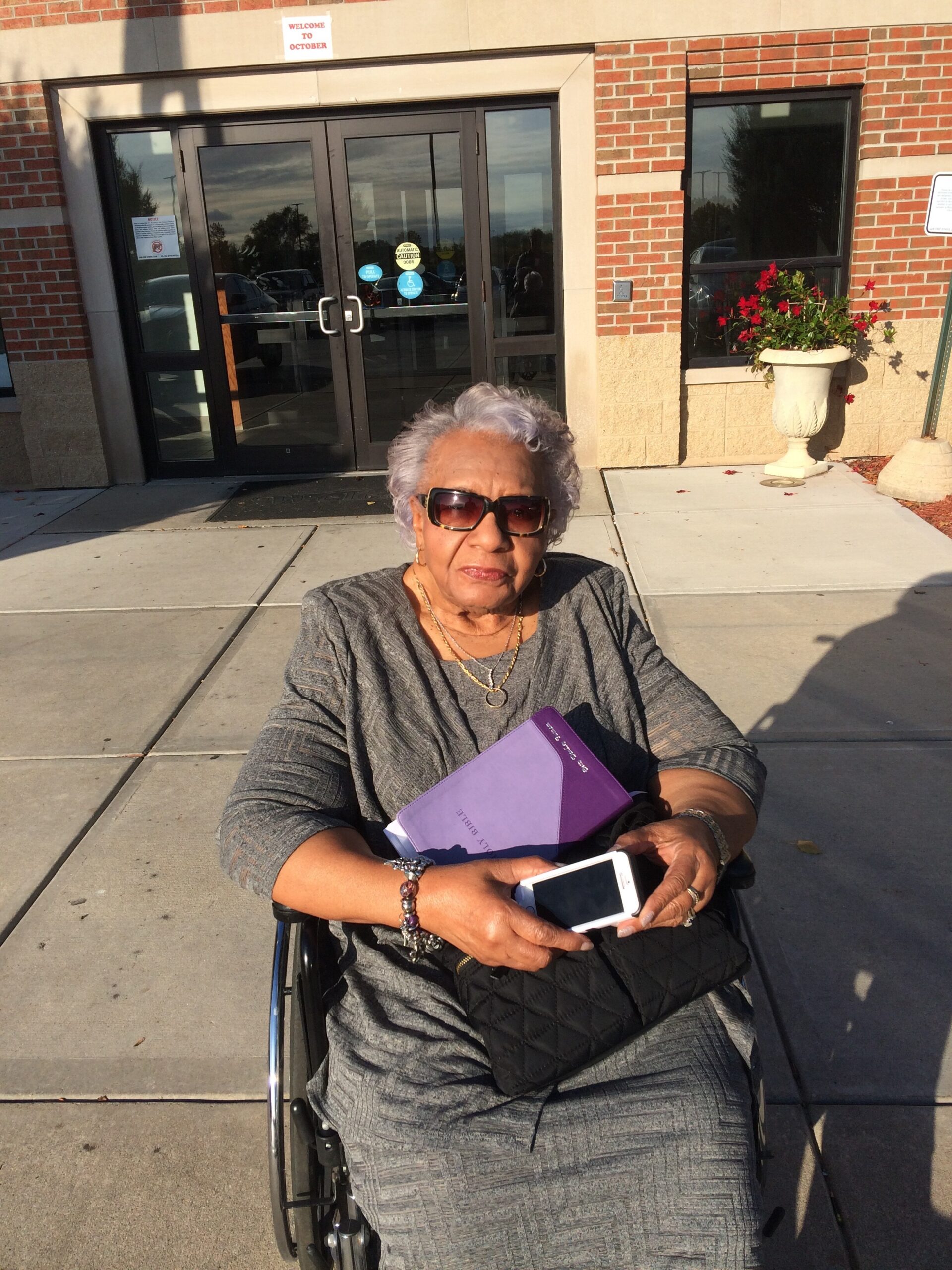 Older woman in a wheelchair holding a purple notebook and a smartphone outside a brick building with glass doors.