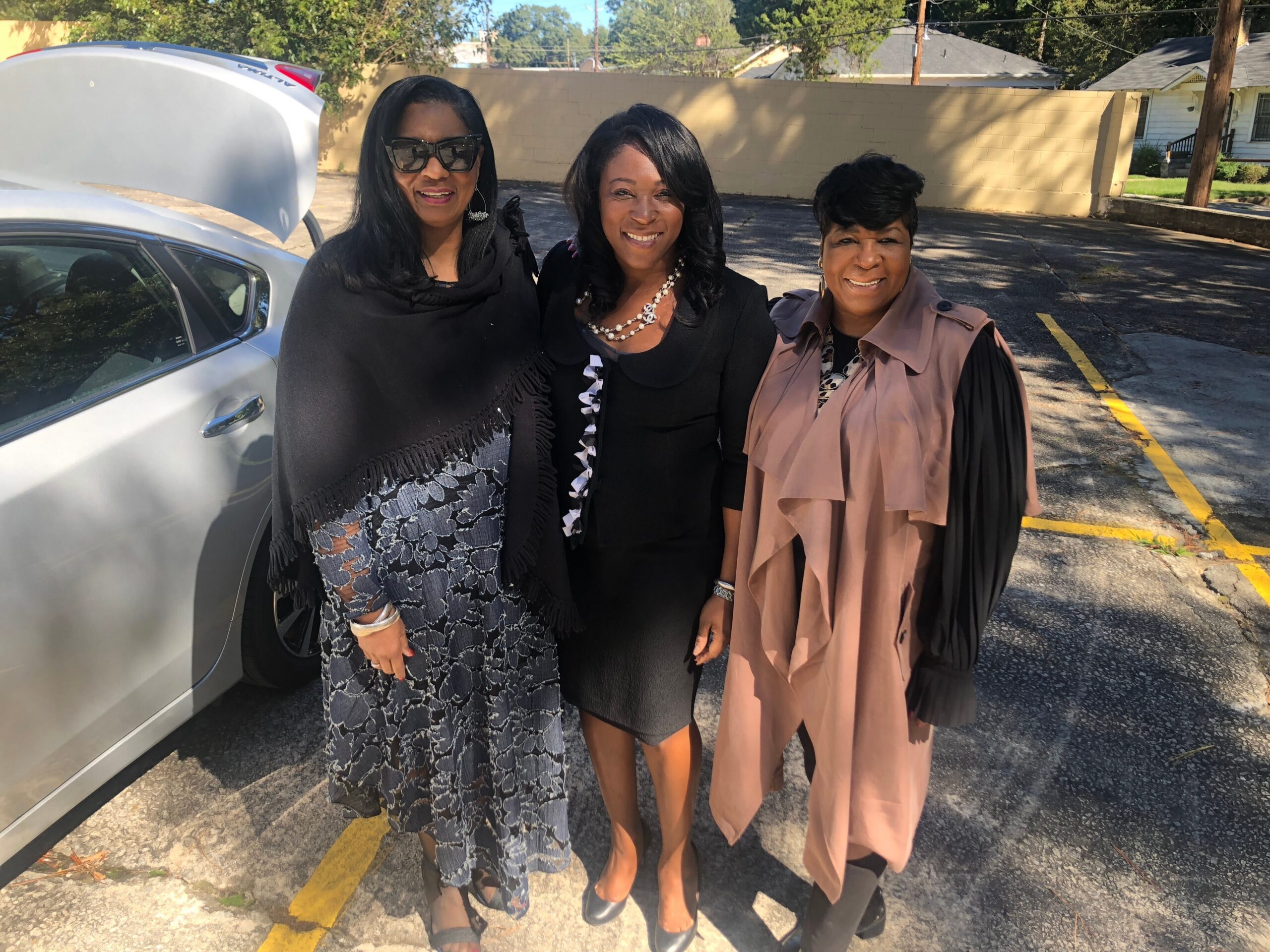 Three women in stylish outfits posing together outdoors in a parking lot beside a white car with its trunk open.