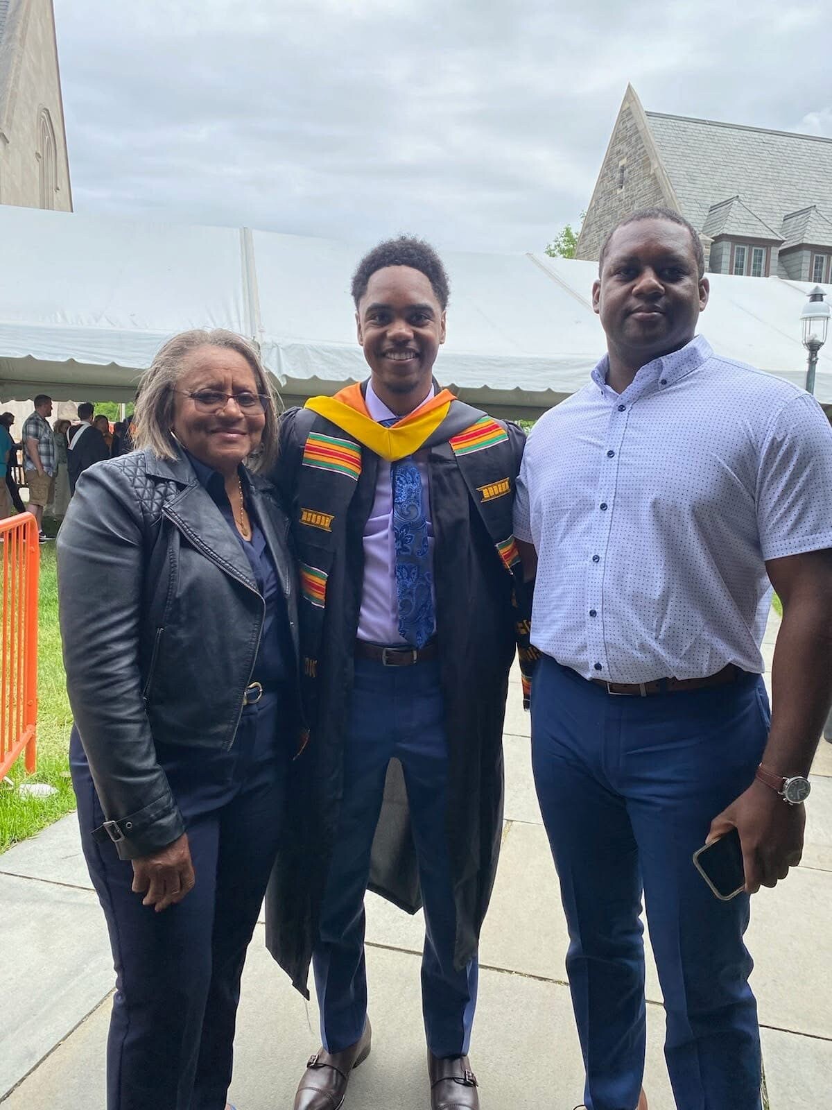 Graduate in cap and gown with a colorful sash posing with an older woman and a man outdoors at a ceremony under a white tent.