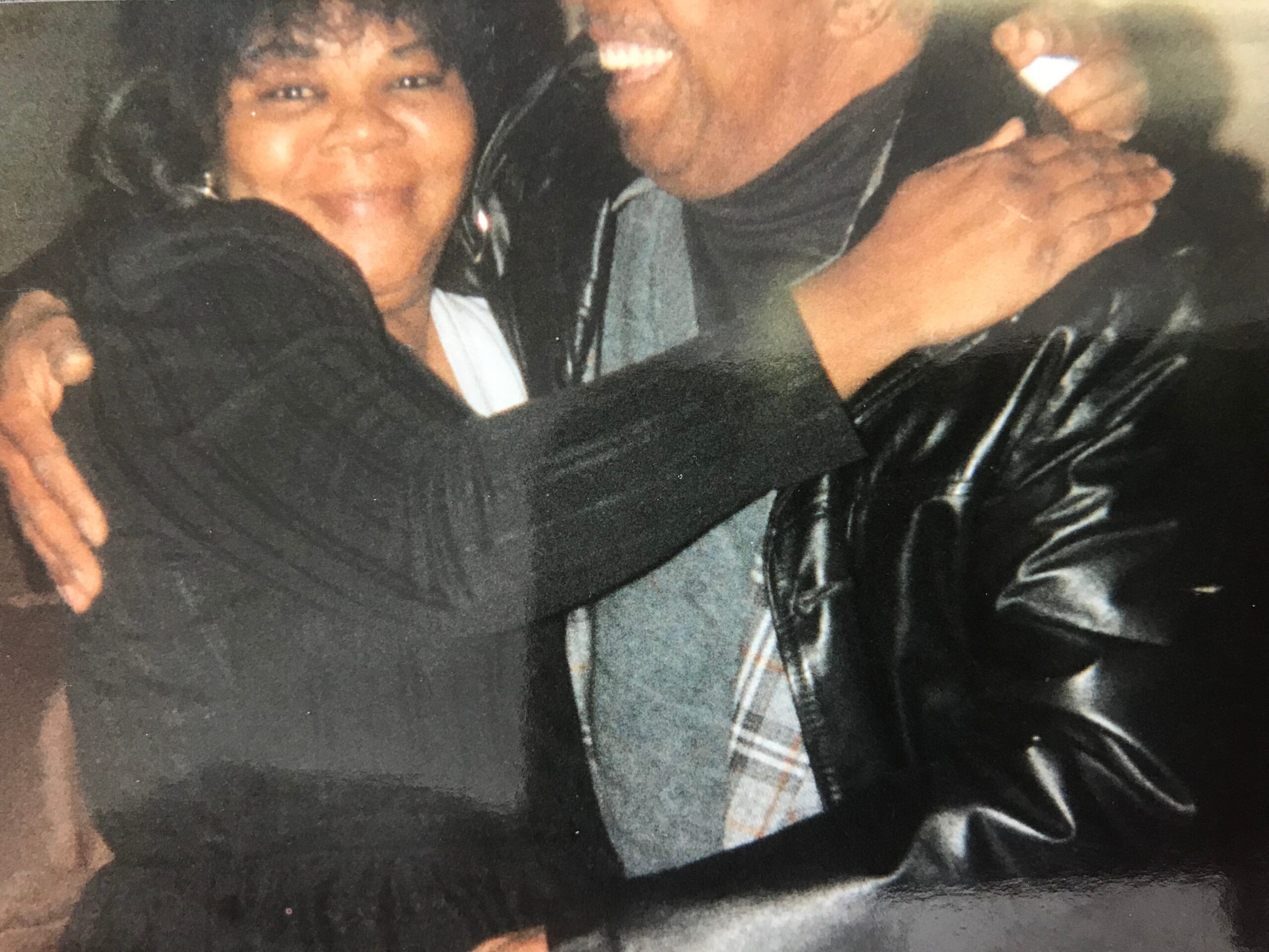Two people embrace in a close hug, smiling, wearing dark clothing and a plaid tie showing between them.