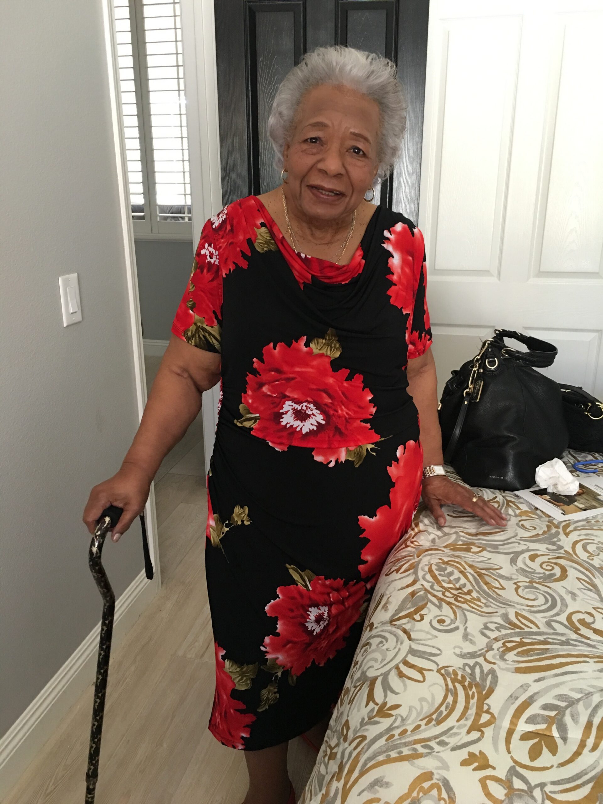 Elderly woman with short gray hair wearing a red and black floral dress, standing with a cane in a bedroom by a bed and a black door behind her.