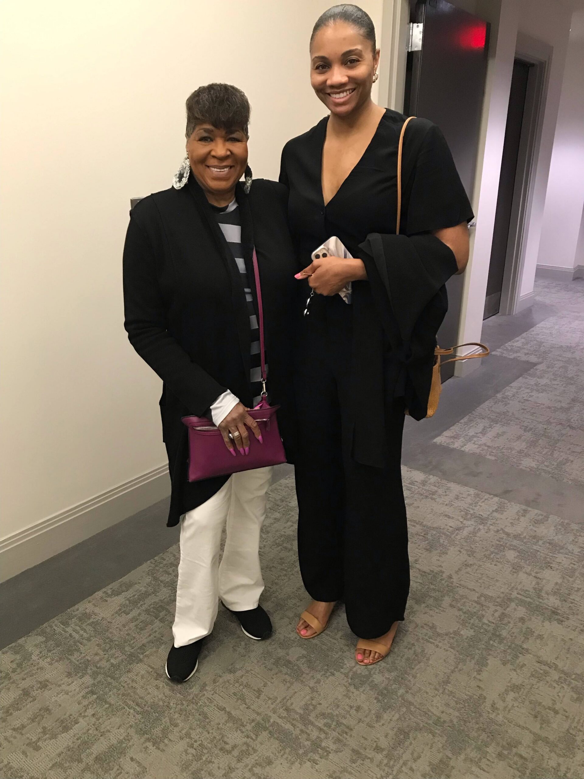 Two women posing in a hallway, both wearing black outfits; one carries a pink purse, the other holds a phone and wears a tan shoulder bag (smiling).