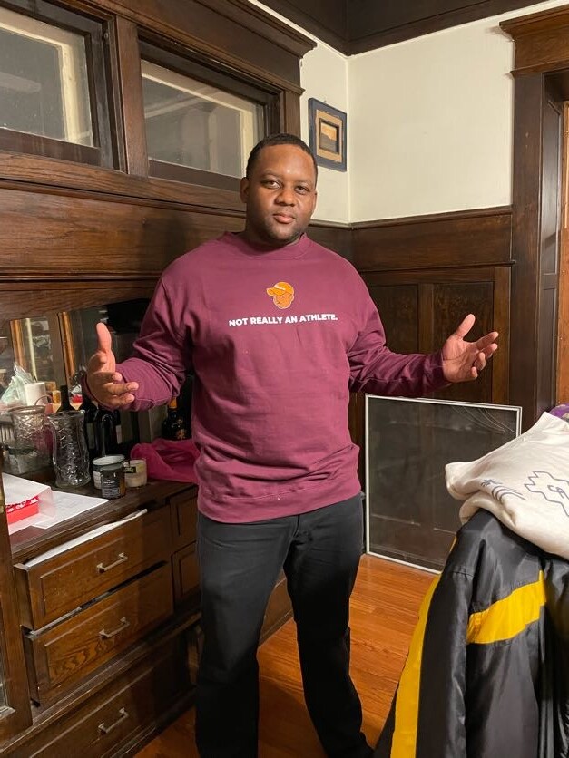 Man indoors in a maroon sweatshirt reading 'NOT REALLY AN ATHLETE,' gesturing with outstretched hands in a wood-paneled room.