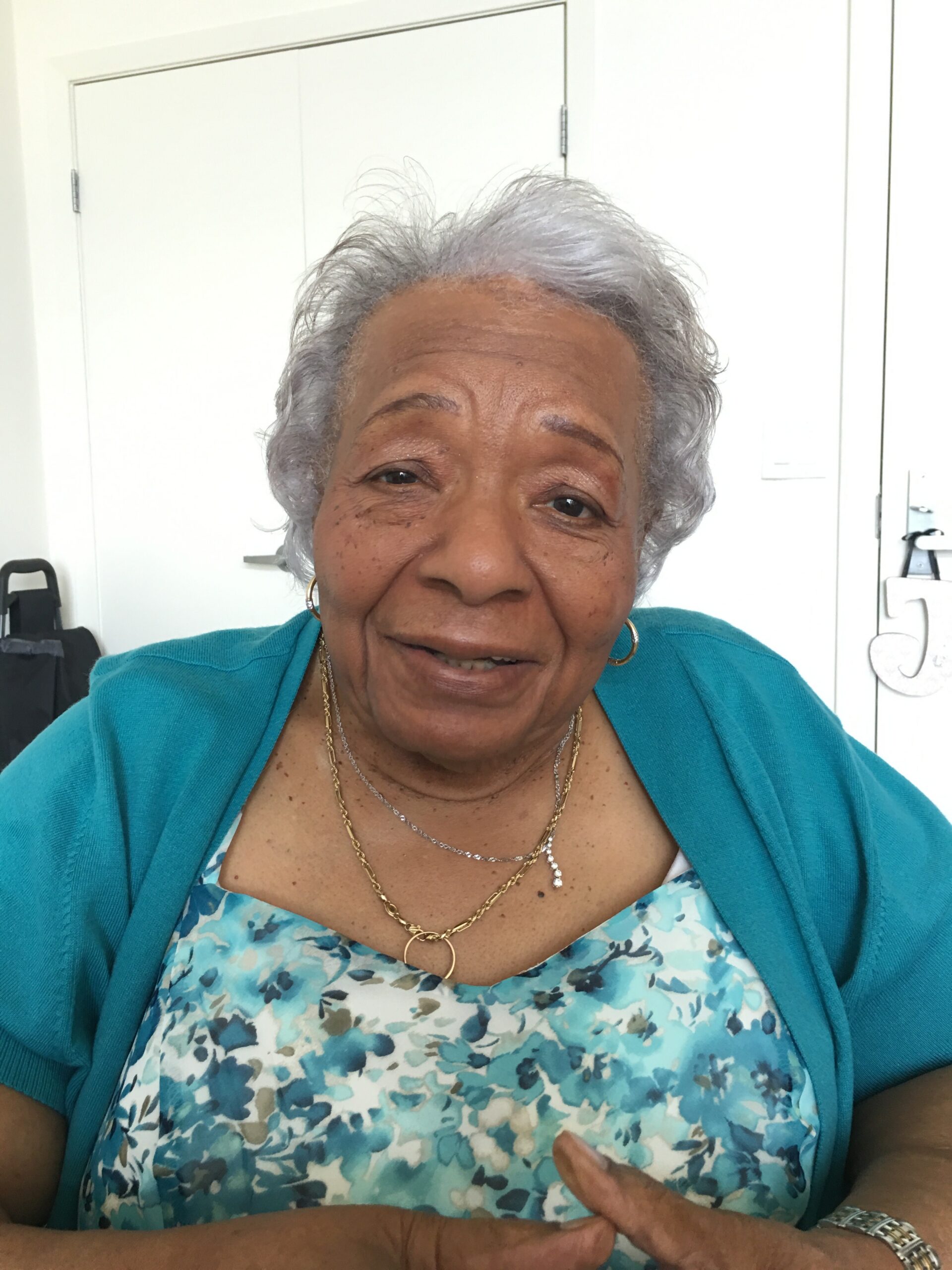 Smiling elderly woman with short gray hair wearing a turquoise cardigan, seated indoors in front of white doors.