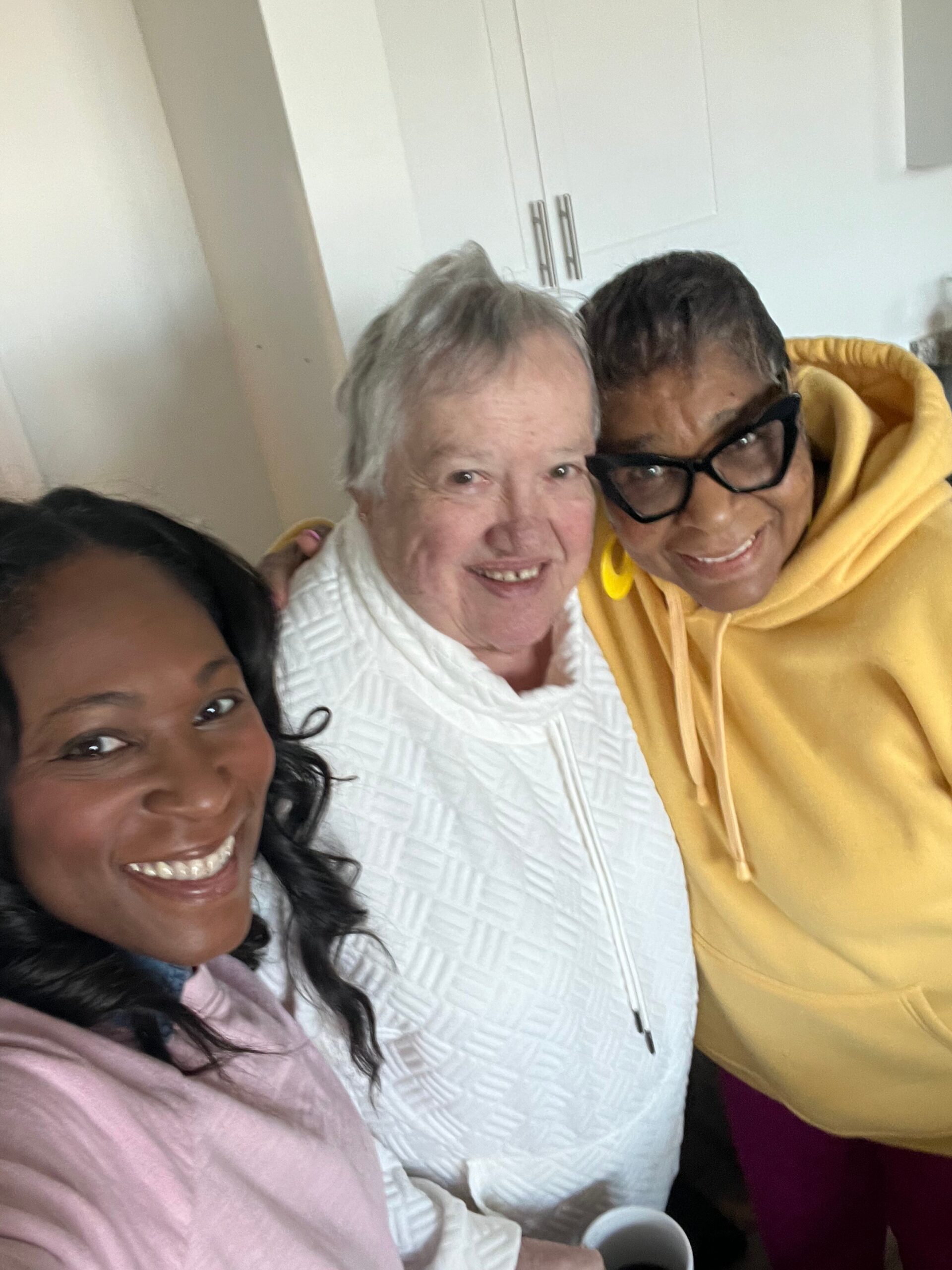 Three women taking a close, cheerful selfie indoors, all smiling at the camera.
