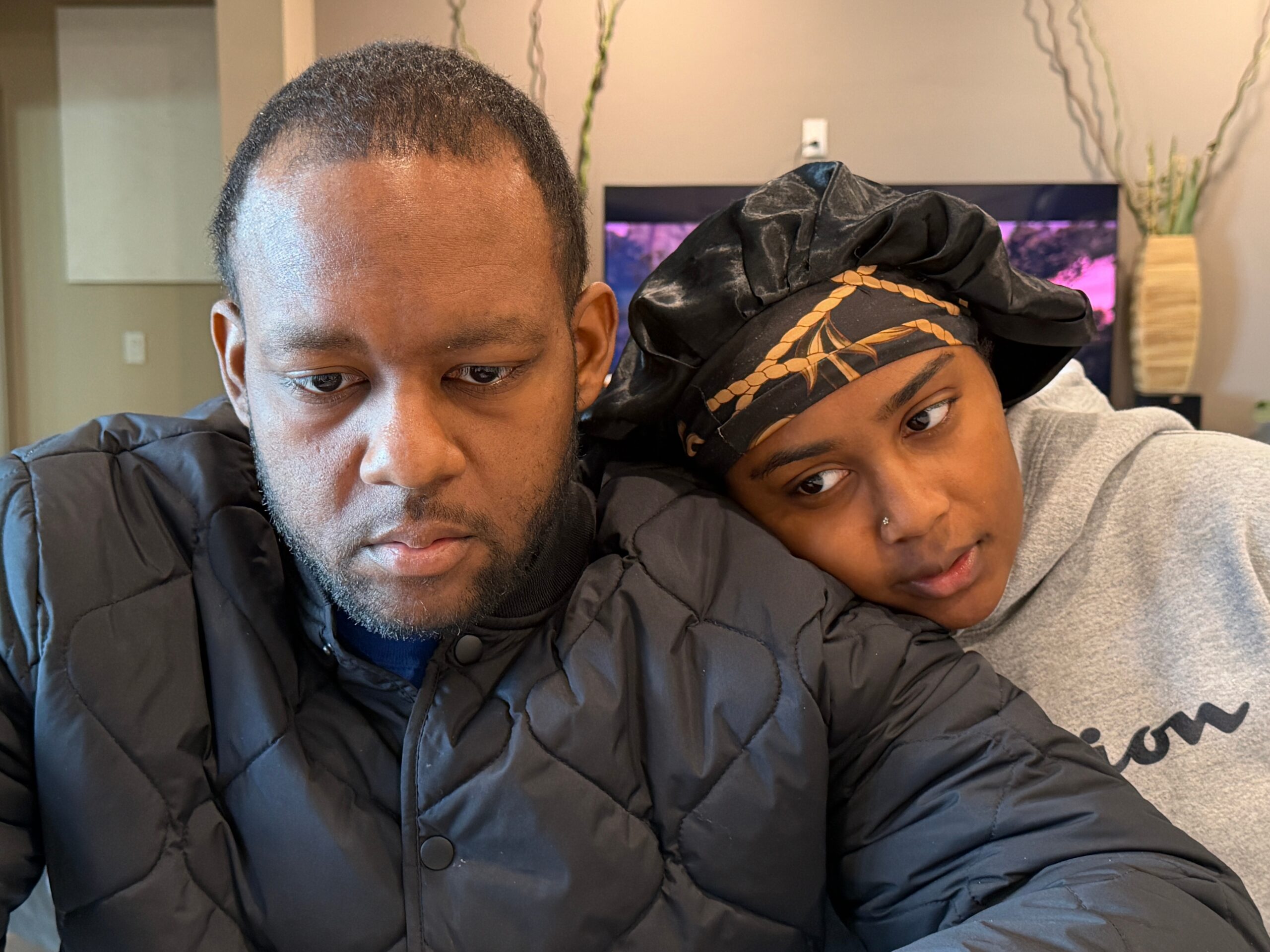 Two people lean their heads together, resting on a couch; both wear dark jackets, and the person on the right wears a black patterned headscarf with orange detailing.