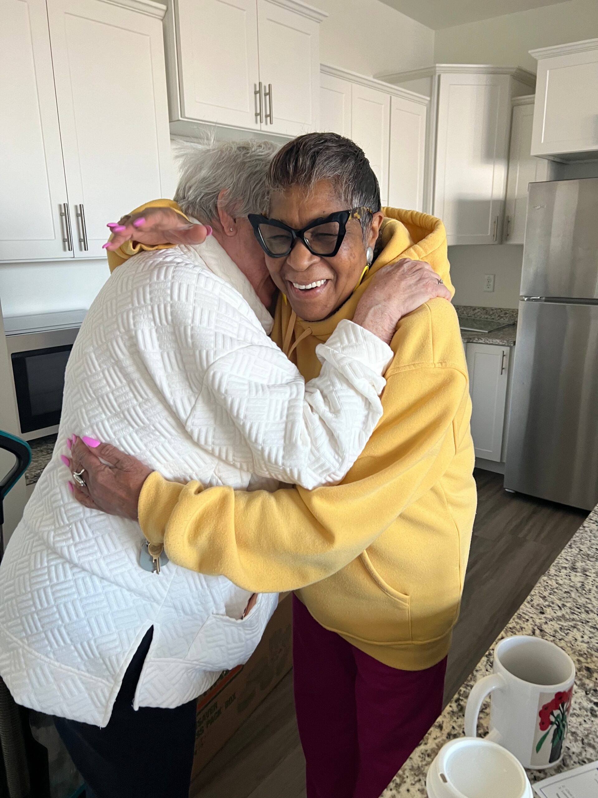 Two elderly women hug in a bright kitchen, smiling; one in a white quilted jacket, the other in a yellow hoodie.