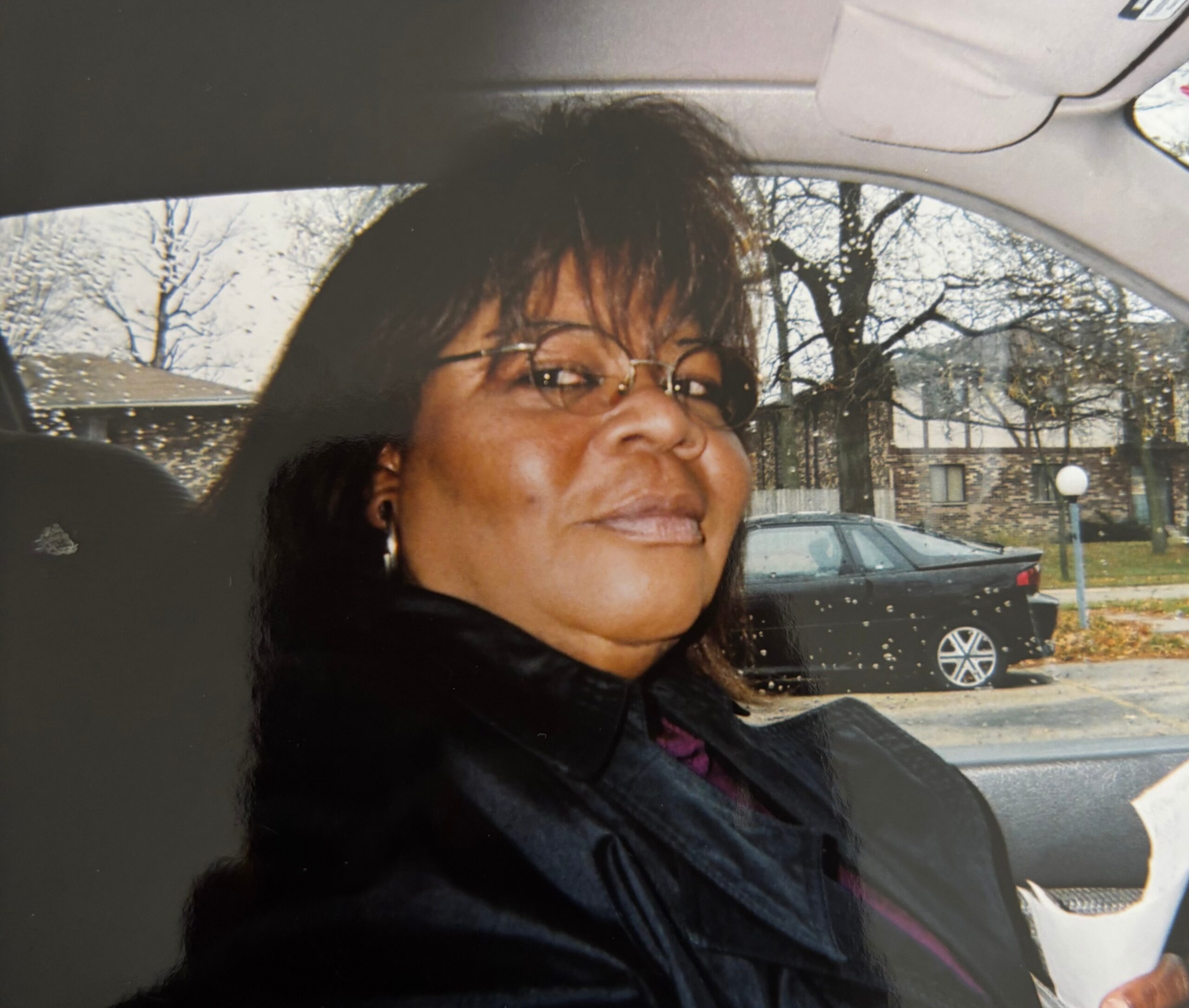 Close-up of a woman wearing glasses inside a car, with trees and houses visible through the window outside.