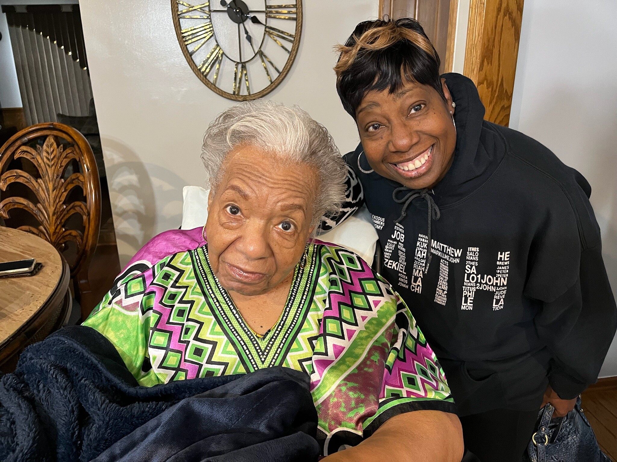 Two women indoors, one elderly in a bright geometric-patterned dress smiling beside a younger woman in a black hoodie.