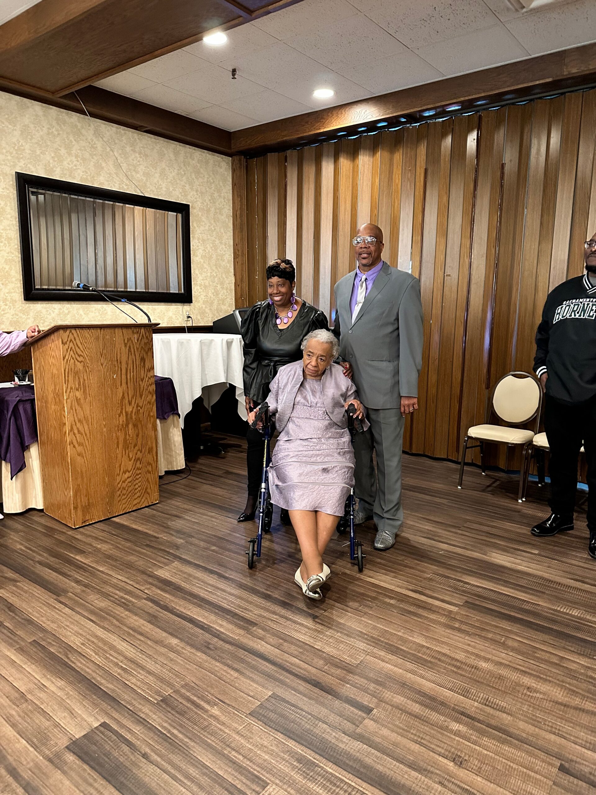 Elderly woman in a lavender dress sits in a wheelchair, flanked by a smiling couple at a formal event in a wood-paneled room.