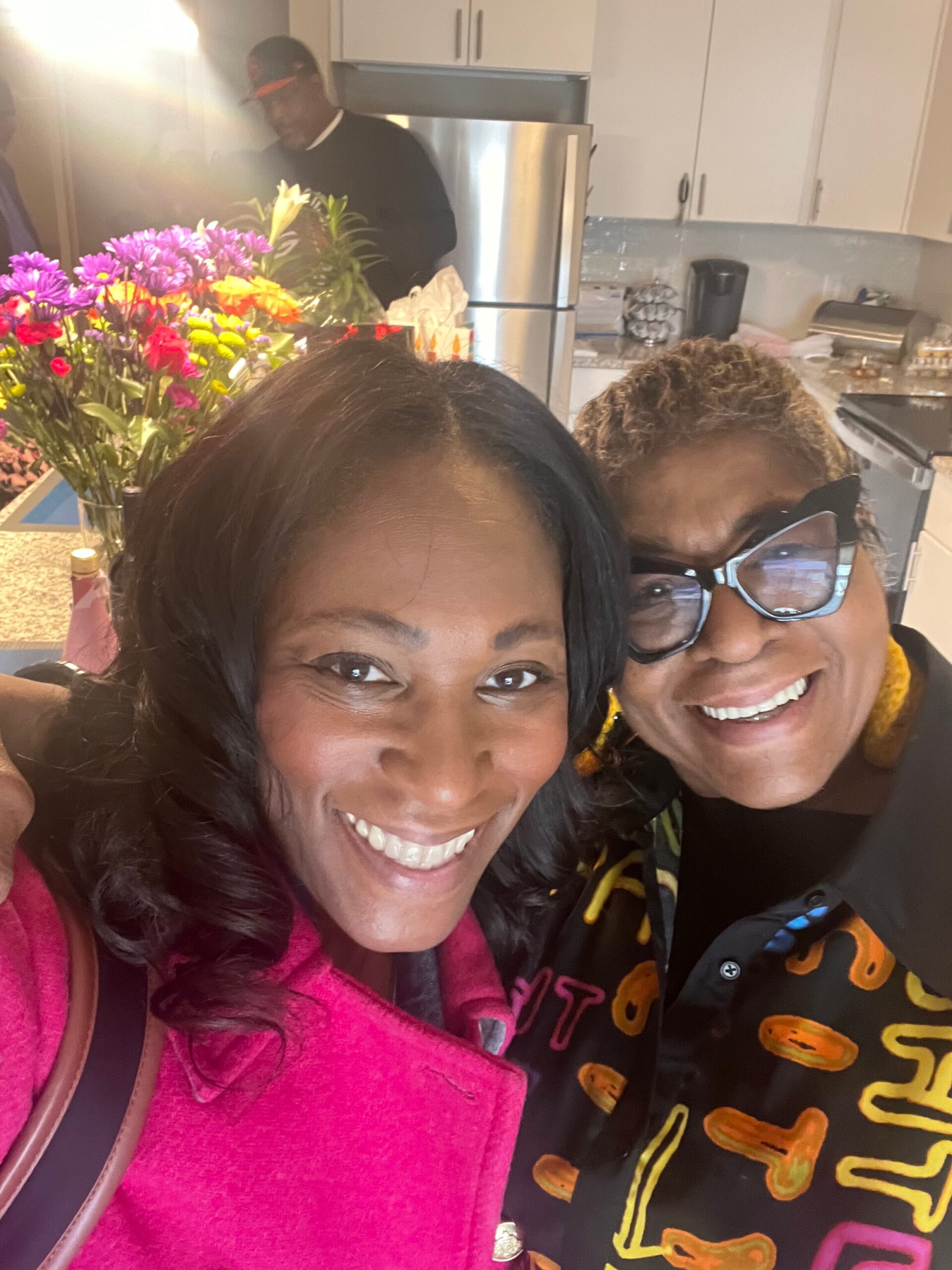 Two women smiling for a selfie in a bright kitchen, with colorful flowers on the counter behind them and a man near the refrigerator in the background.