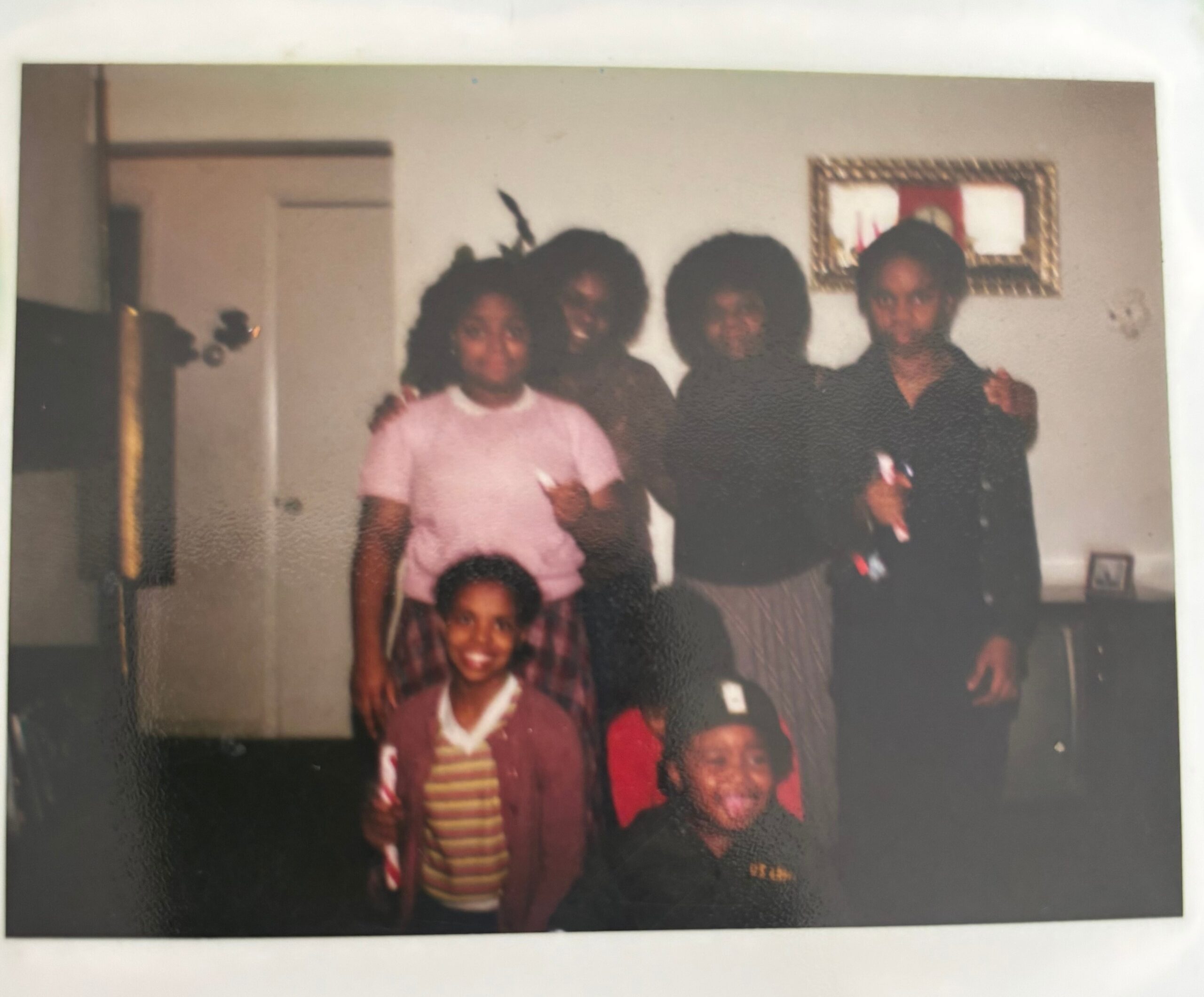 Group of people posing together indoors for a casual photo, several adults and children smiling at the camera, a framed picture on the wall behind them.