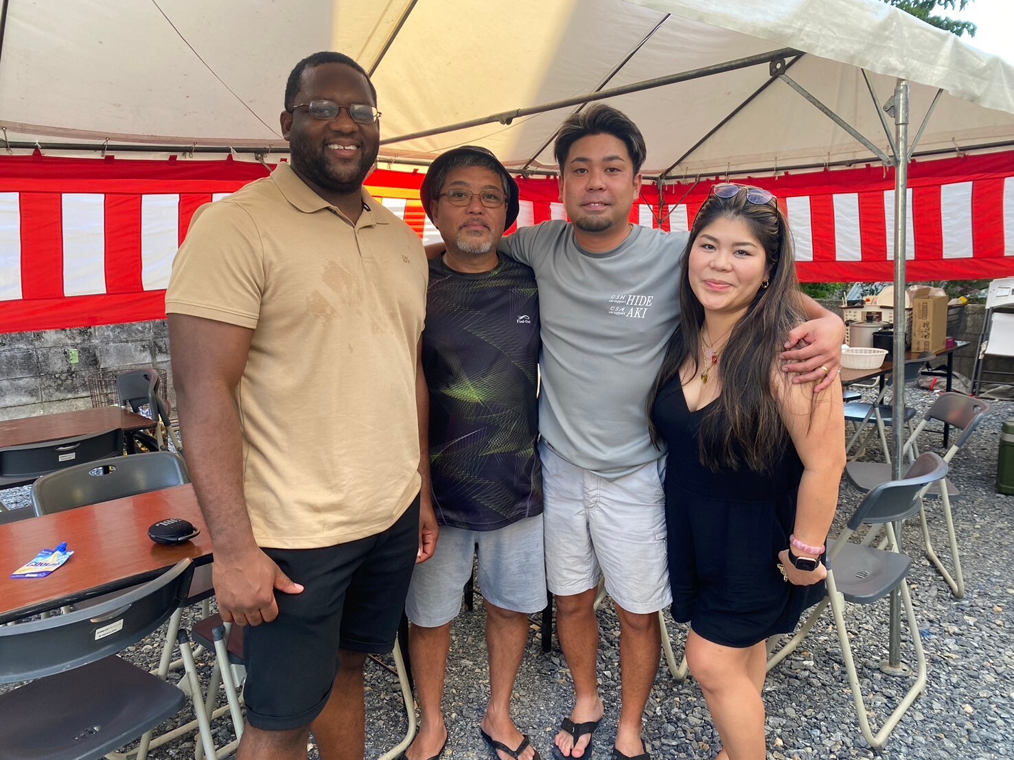 Four friends posing together under a white canopy with red-and-white striped banners, smiling at the camera, outdoors at a casual event.