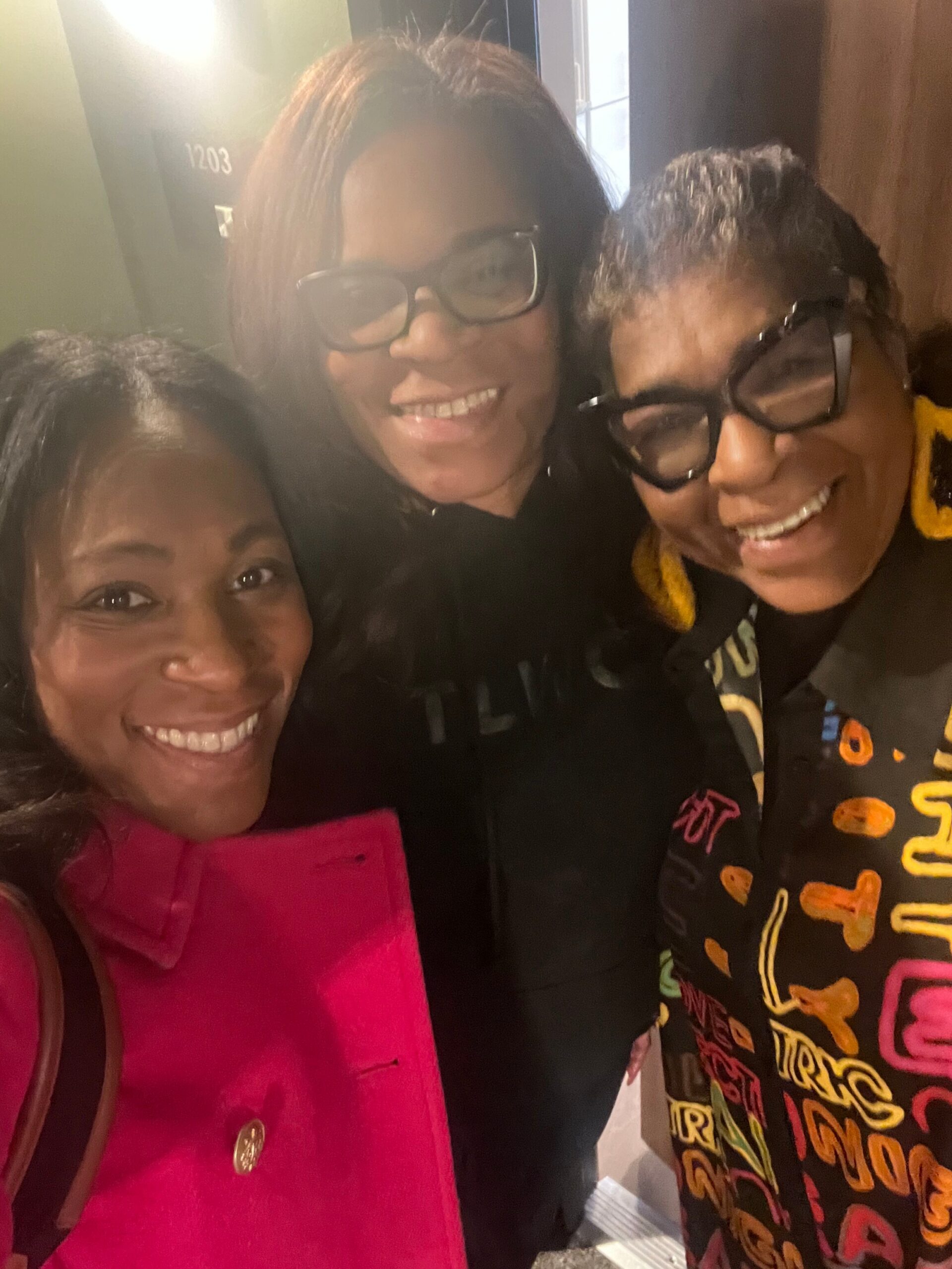 Three friends posing for a close-up selfie indoors, smiling at the camera with colorful outfits.