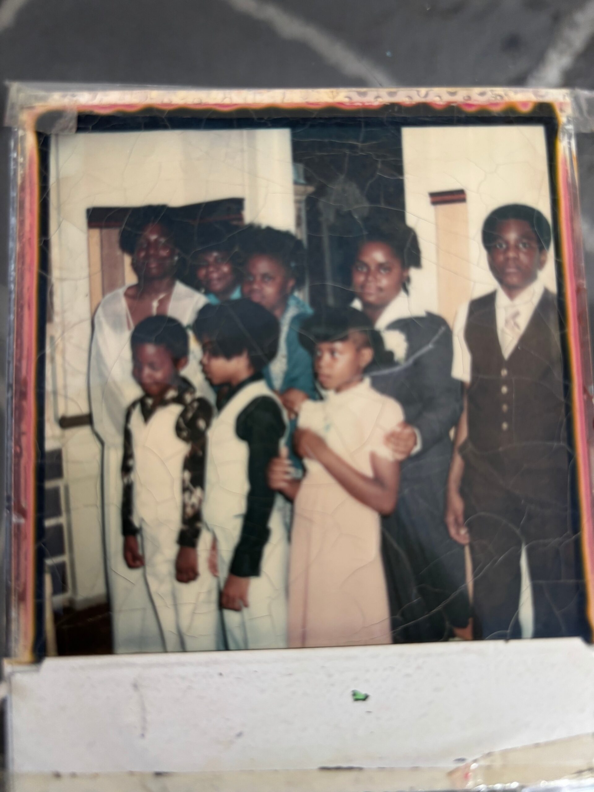 Group of adults and children posing for a family portrait, dressed in formal clothes in a doorway setting, old photo with cracks and wear in the frame.
