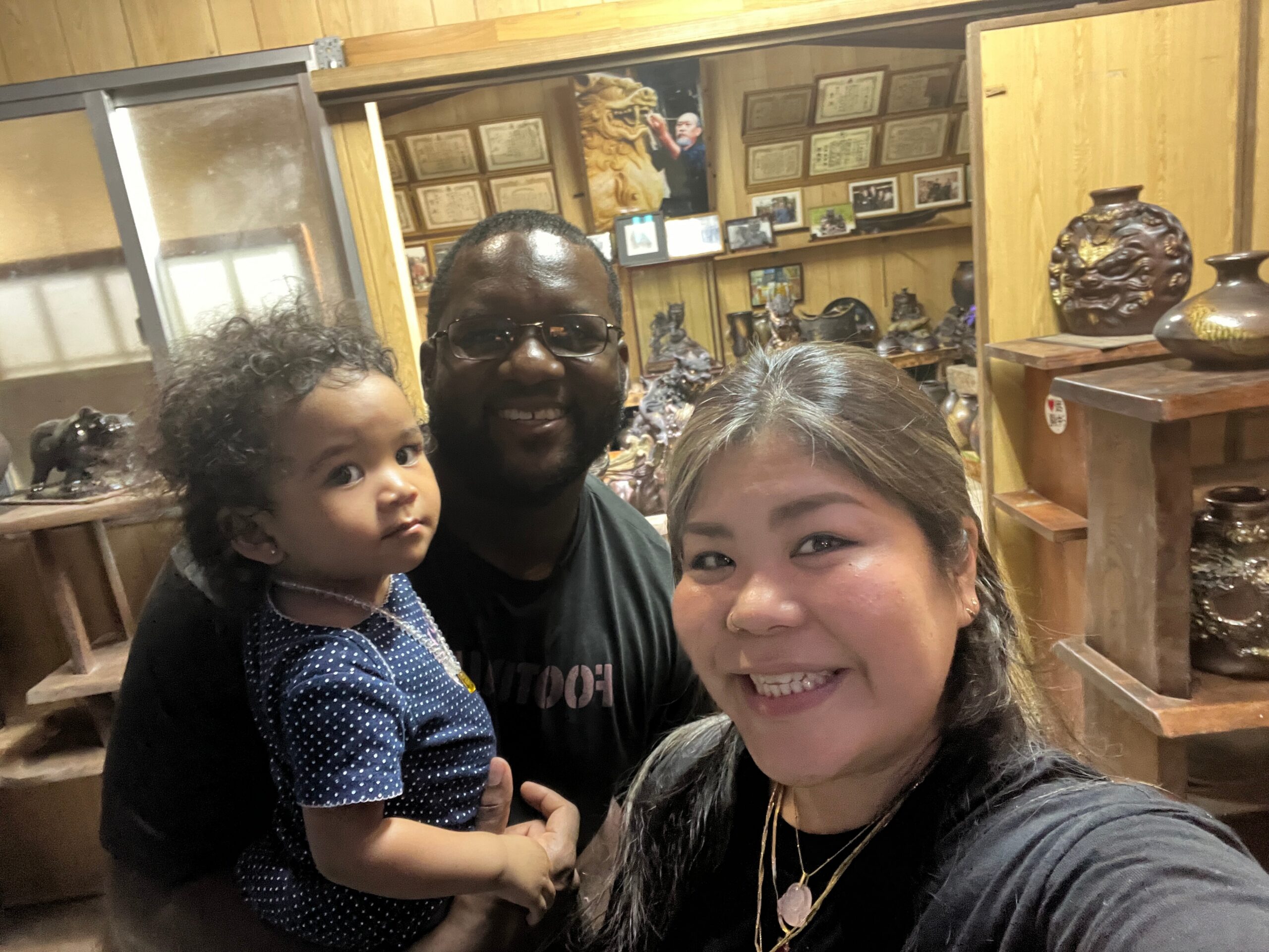 Three people take a close selfie in a wood-paneled room filled with ceramic vases and shelves behind them.