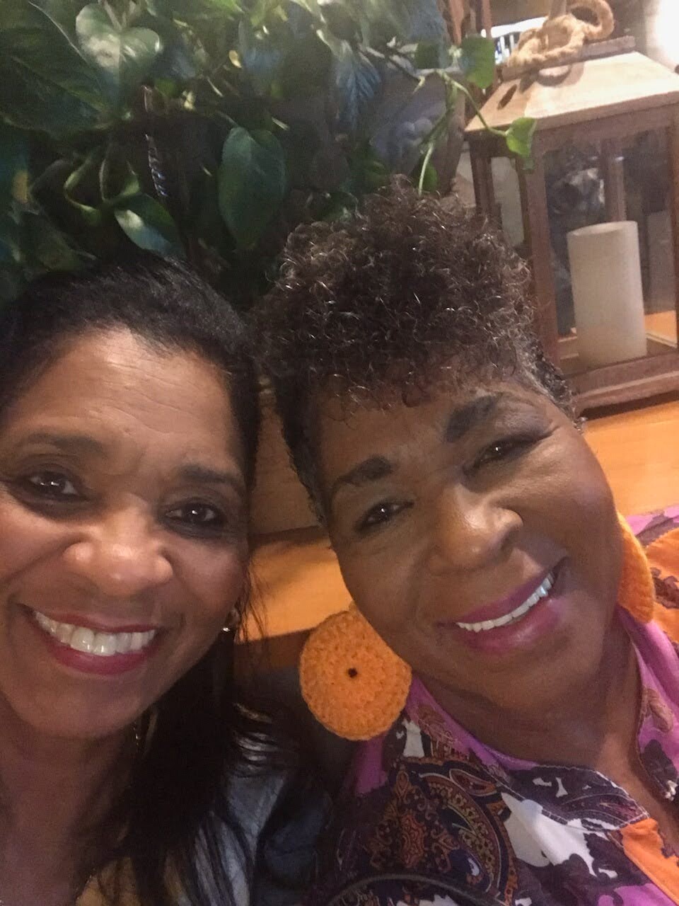 Two women smiling for a selfie indoors, with green plants and a wooden lantern in the background.