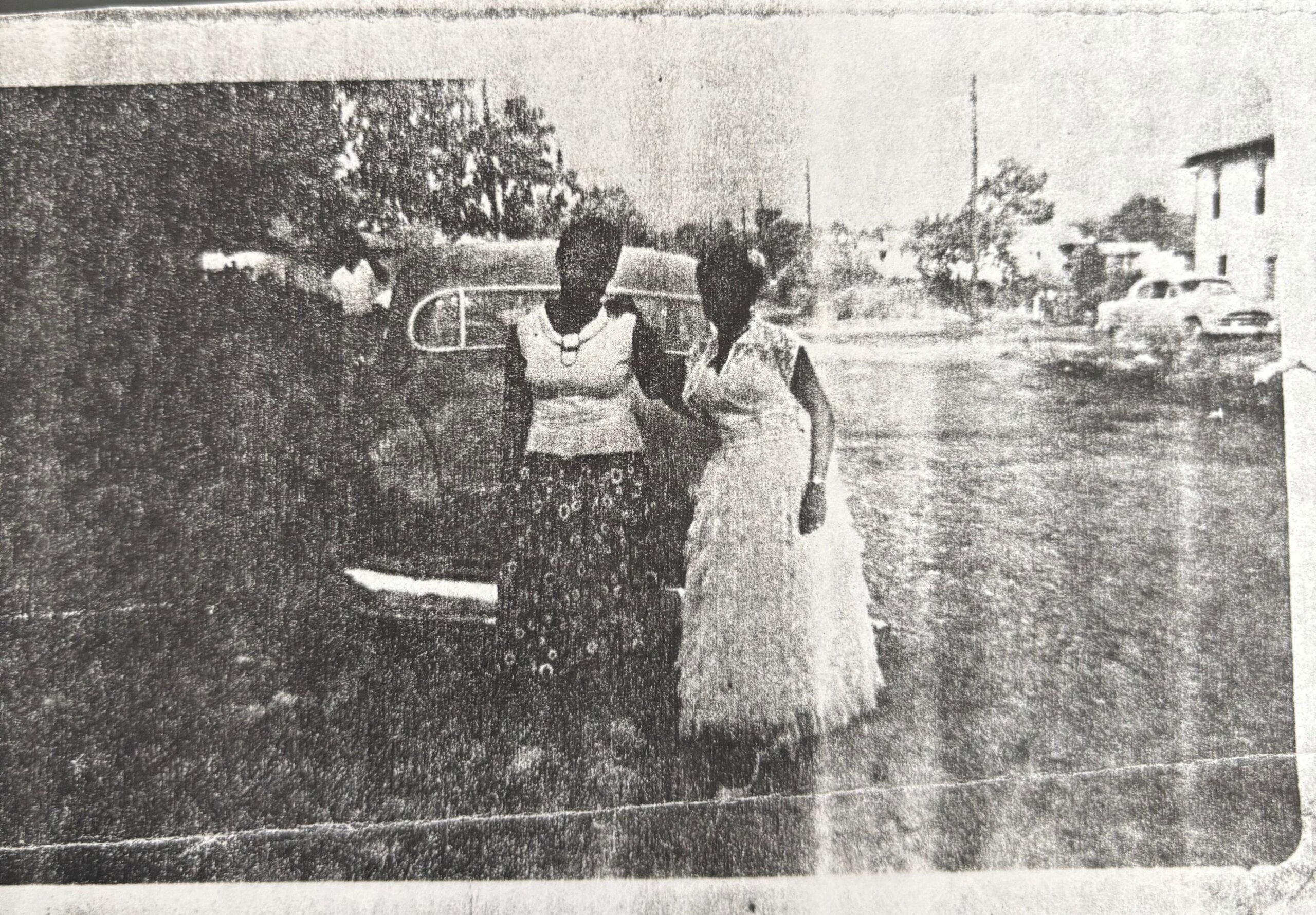 Two women walk together on a street near a parked car and a white building in the background, older photograph style.