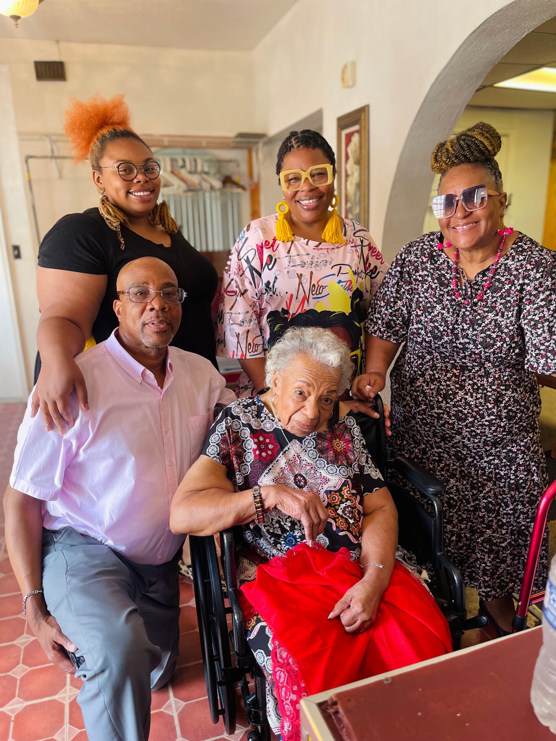 Family gathers for a photo indoors: elderly woman in a wheelchair center, flanked by four adults with bright outfits and smiles.