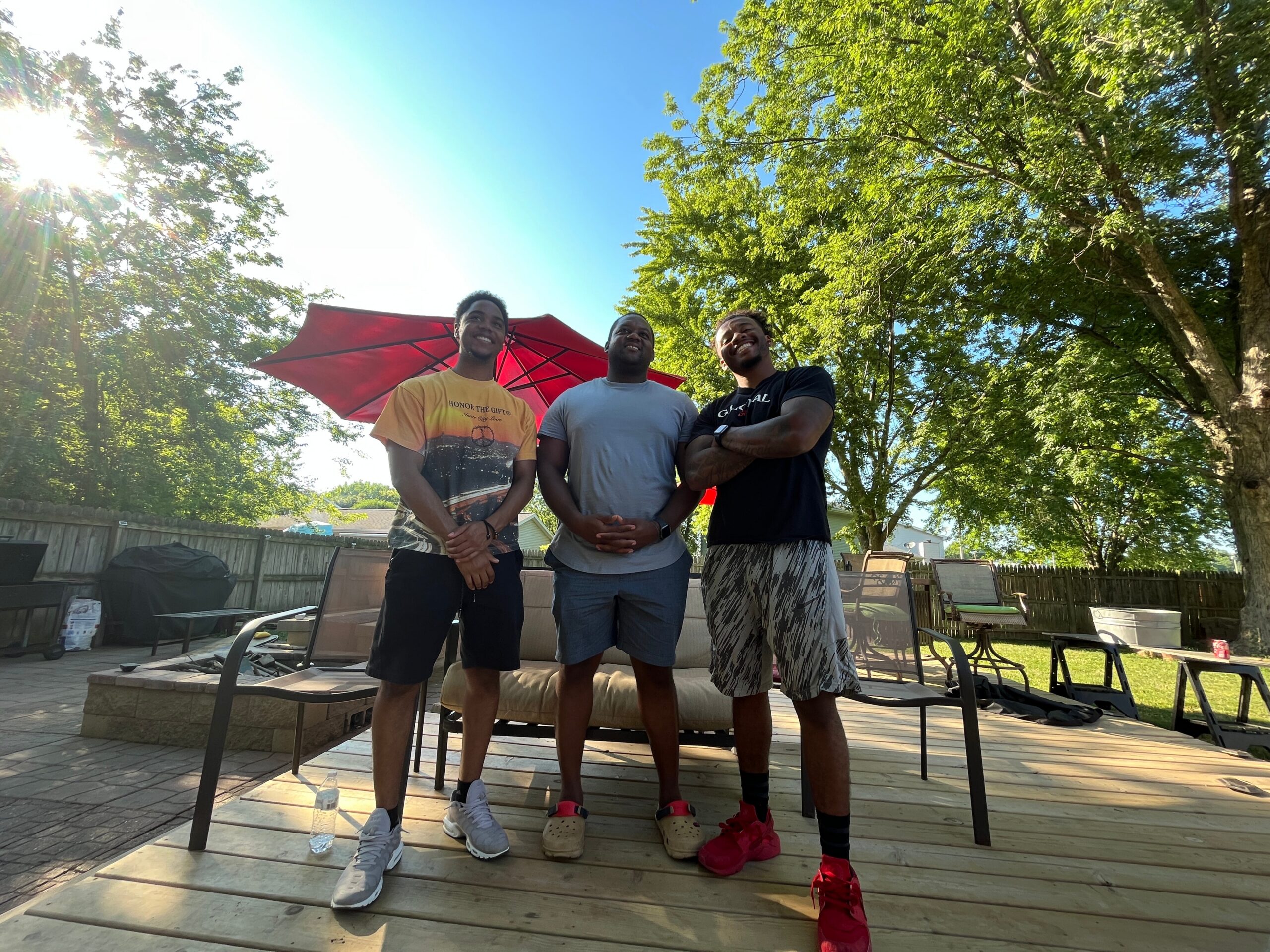 Three men stand together on a wooden backyard deck, smiling, with a red umbrella and sunlit trees in the background.