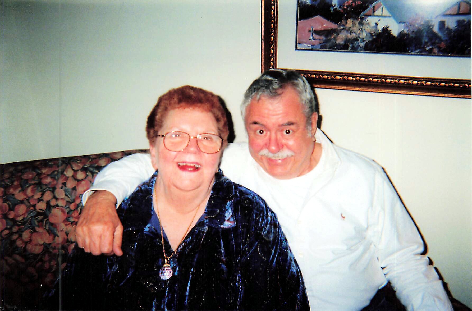 Smiling elderly couple sitting on a floral-patterned couch with arms around each other indoors.