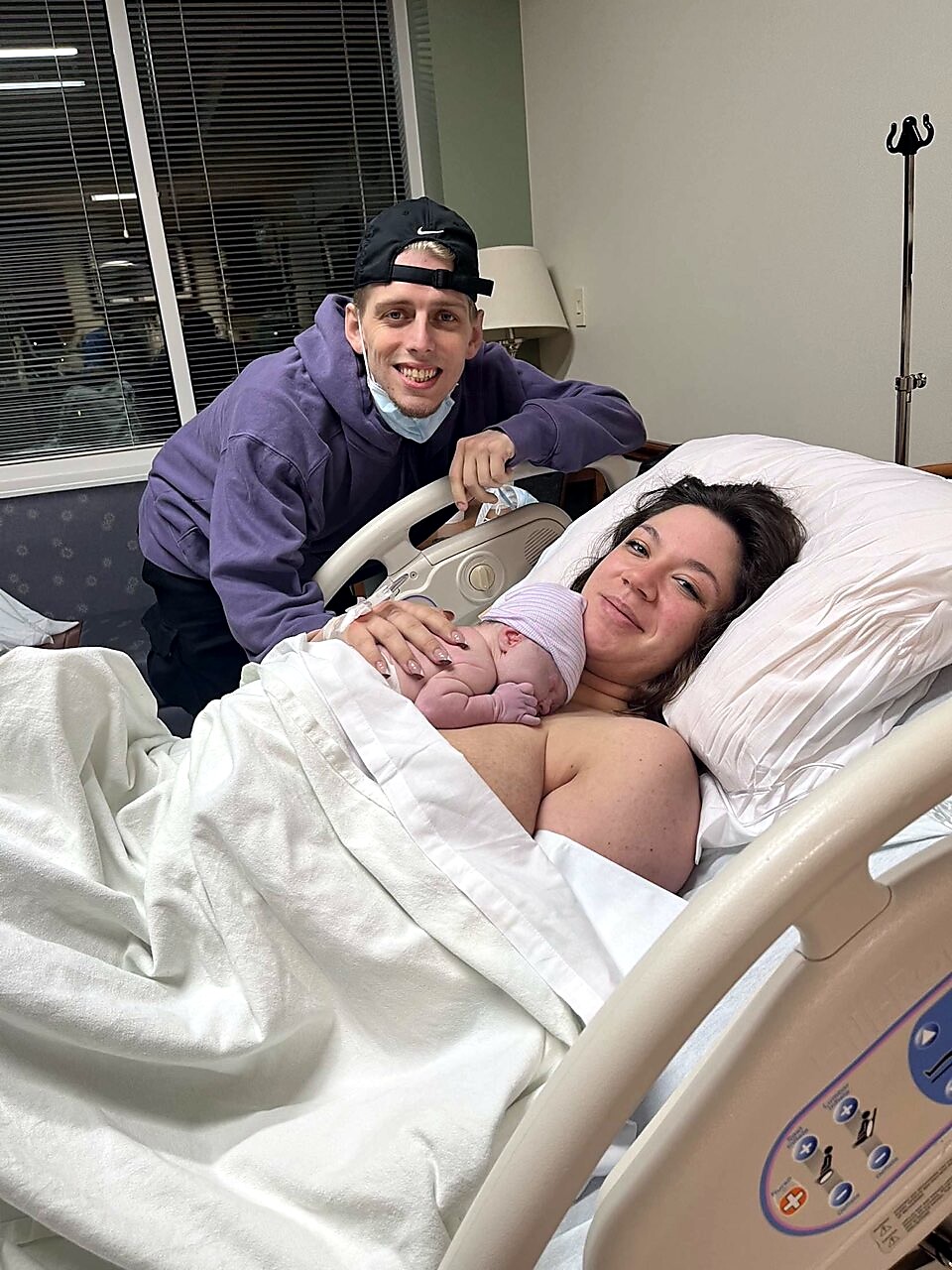 Mother in a hospital bed cradling her newborn baby as a smiling partner in a purple hoodie leans over the railing nearby, in a hospital room with blinds in the background, wearing a mask beneath.
