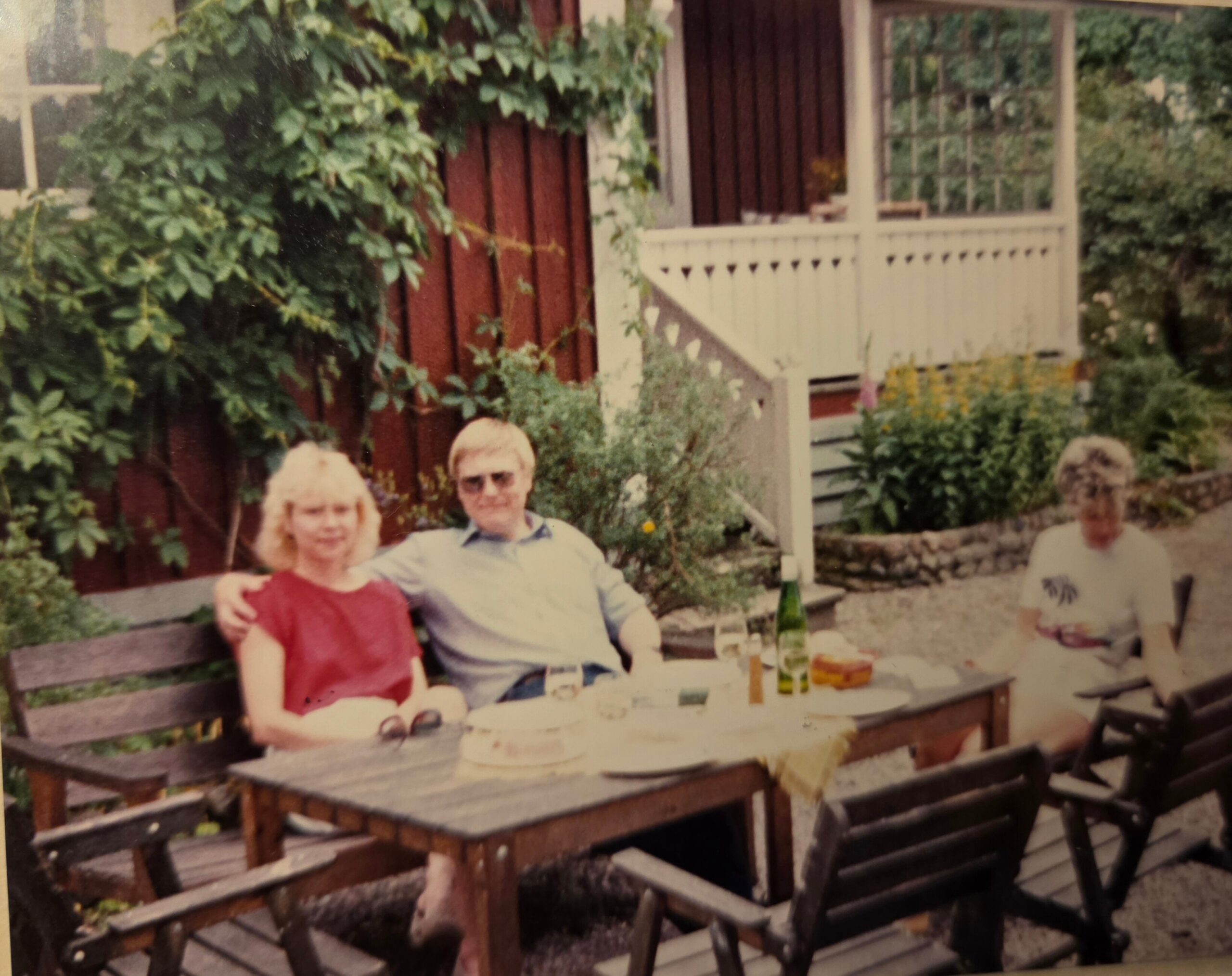 People gathered around an outdoor table in a garden, with drinks and a cake visible on the table, near a house with a porch in the background.