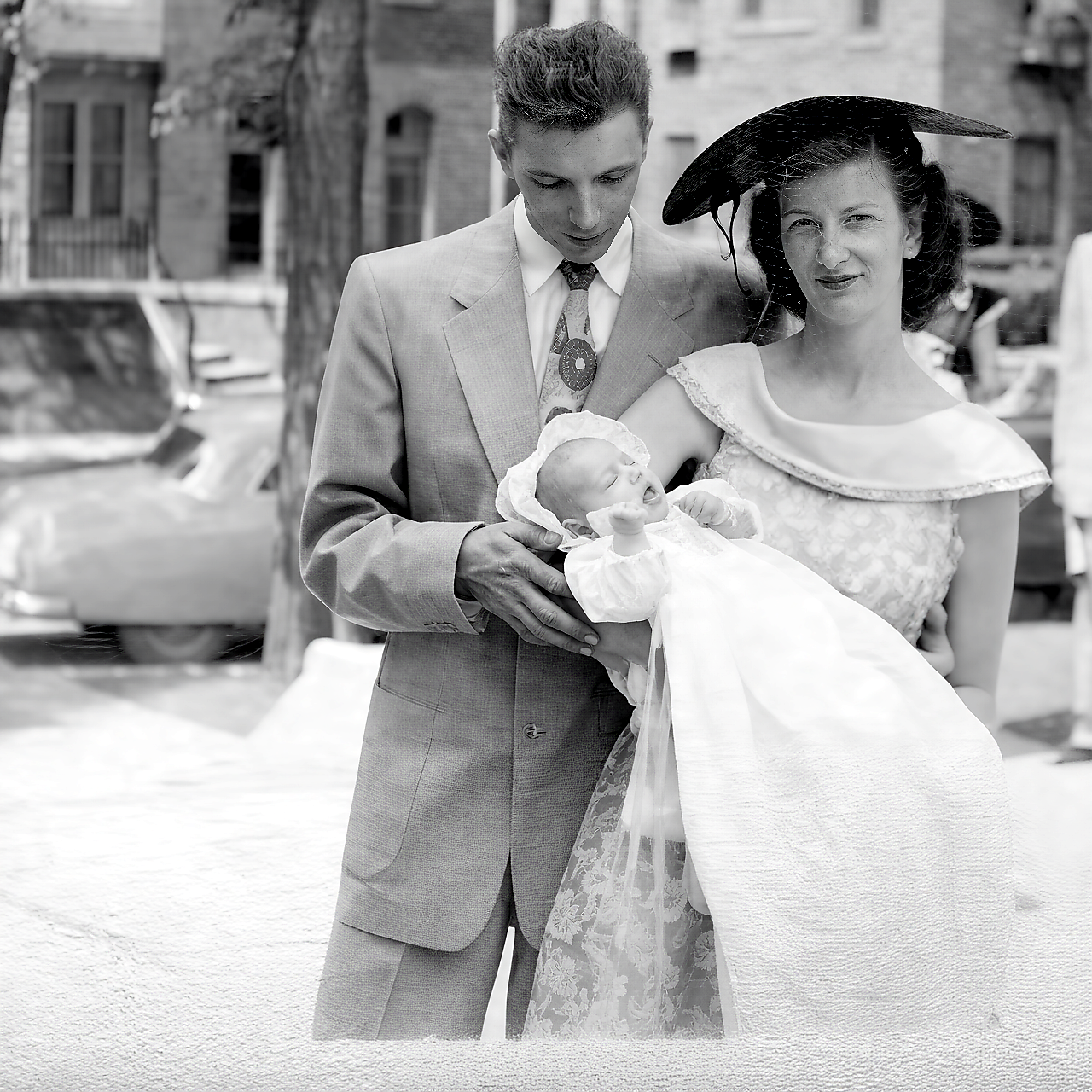 A couple in vintage wedding attire holding a newborn baby in a white gown outdoors, black-and-white photo.