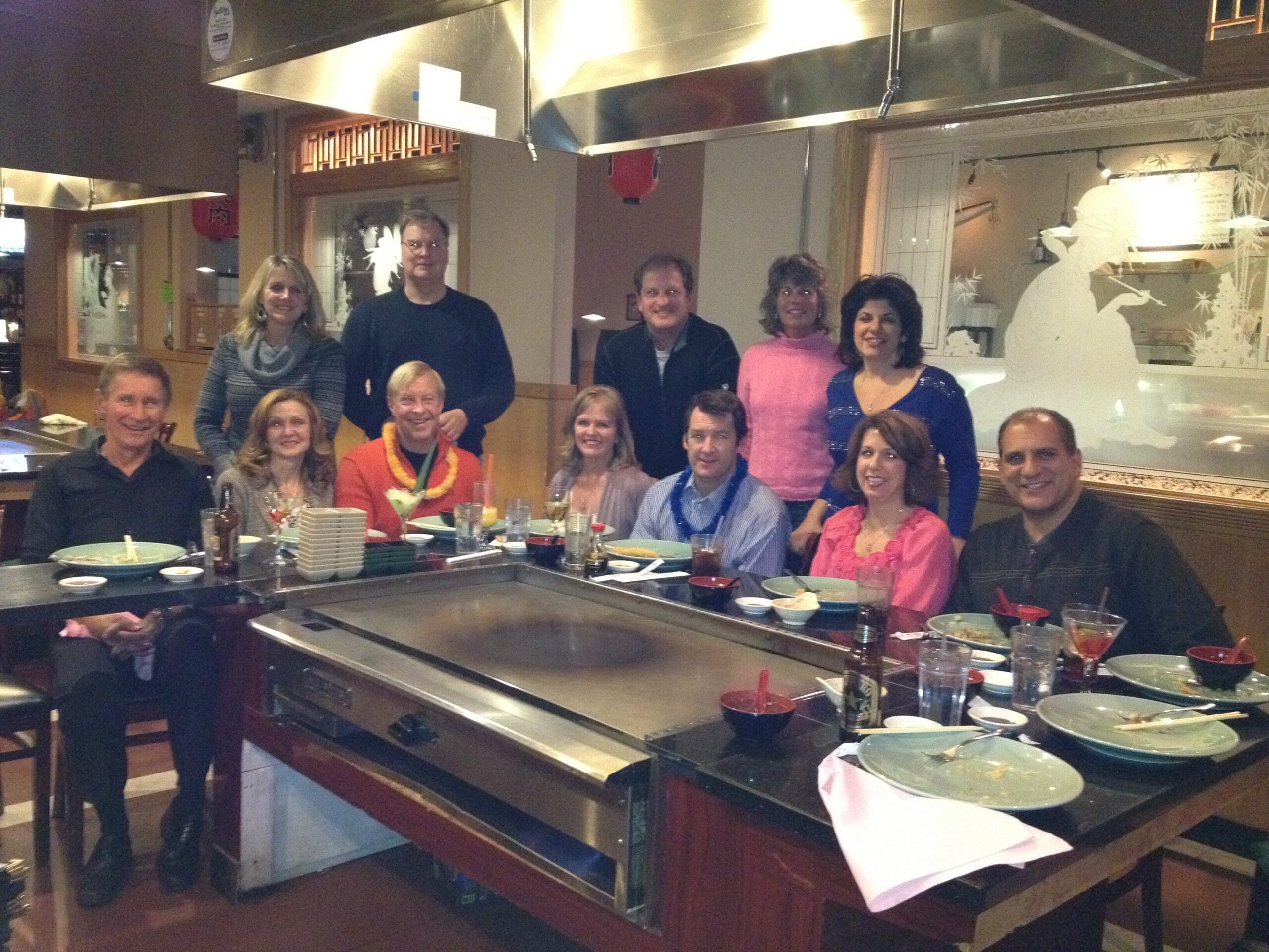 Group of adults posing and smiling around a hibachi grill at a restaurant, with plates and drinks on the table behind them.