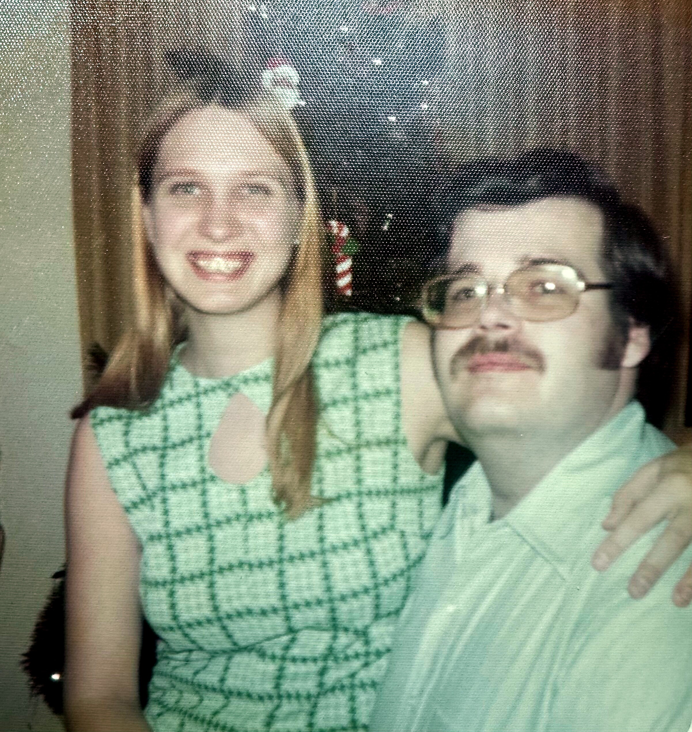 Two friends posing for a selfie: a smiling woman with long light hair wearing a green plaid shirt, and a man with glasses and a green shirt resting his chin on his hand.
