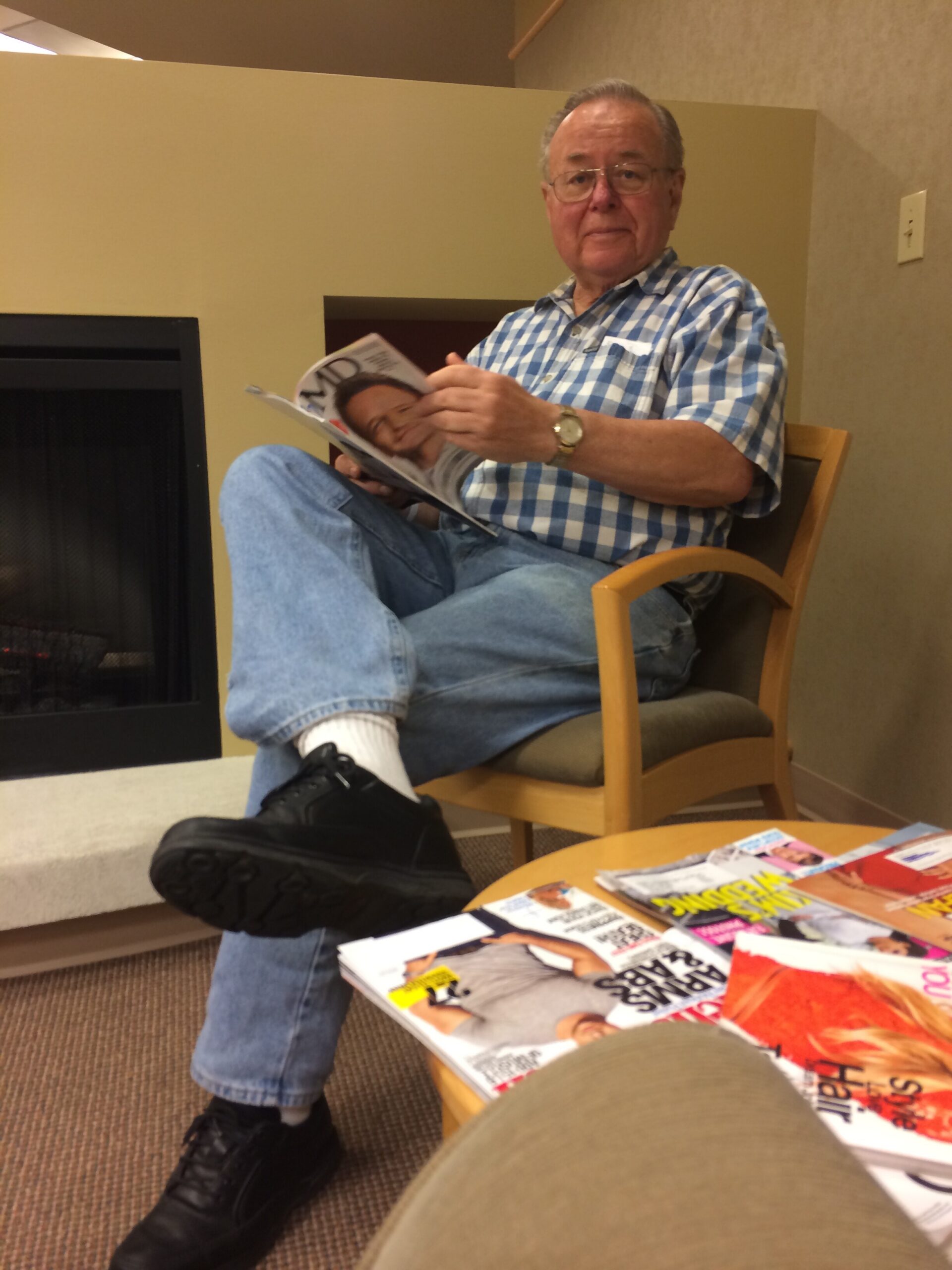Older man in a blue plaid shirt sits in a wooden chair by a fireplace, reading a magazine laid open on his lap.