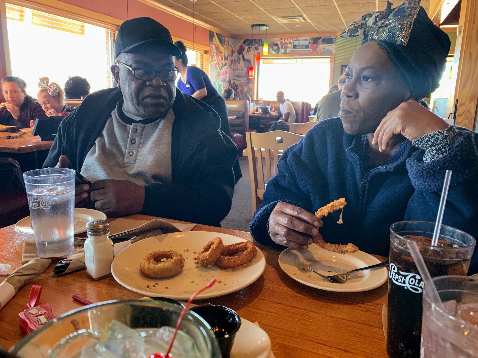 Two people sit at a wooden table in a lively restaurant, sharing onion rings and drinks with a mural-filled background behind them.
