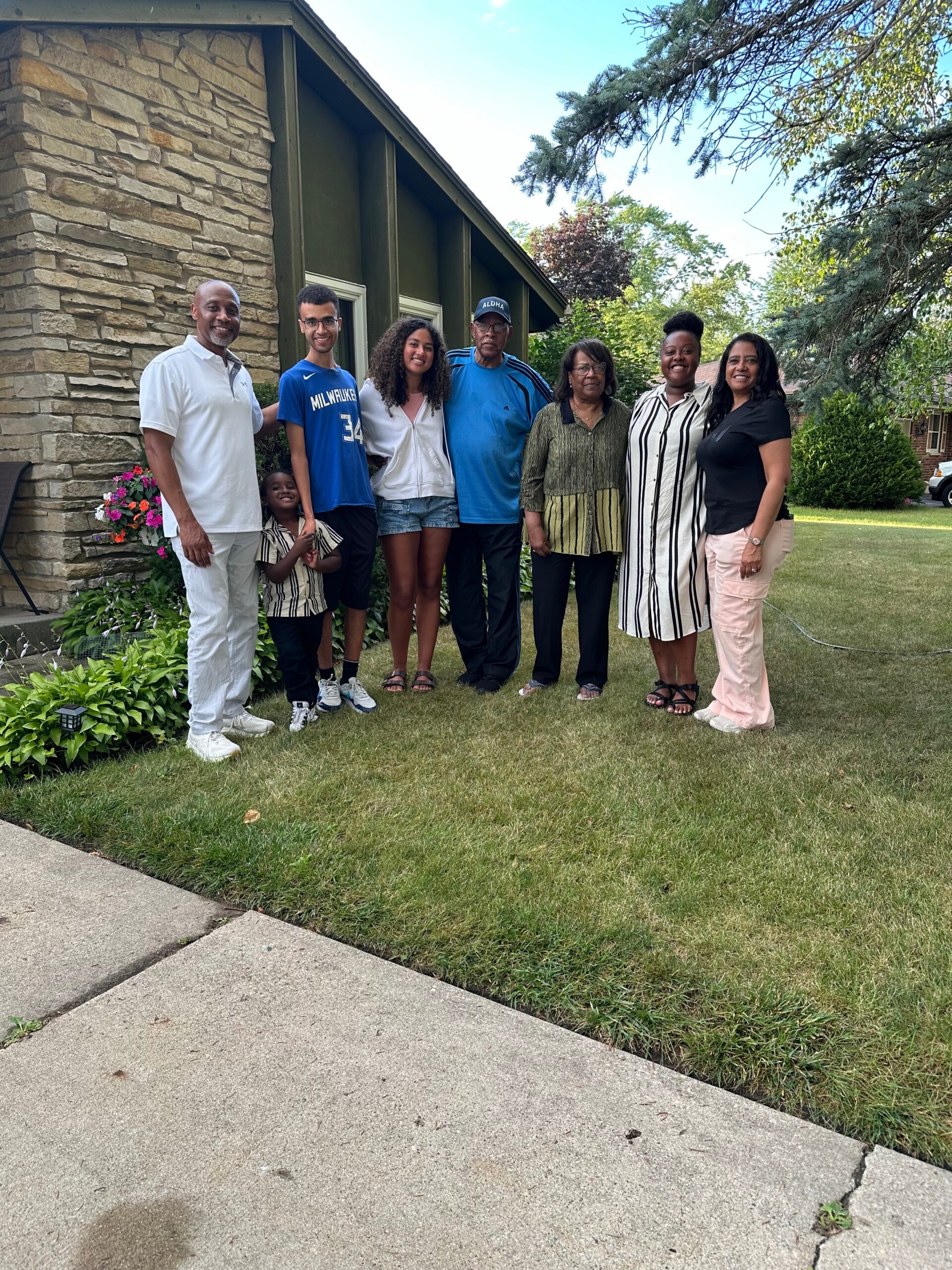 Group of eight people posing together on a front lawn in front of a stone house, smiling for a family photo on a sunny day.